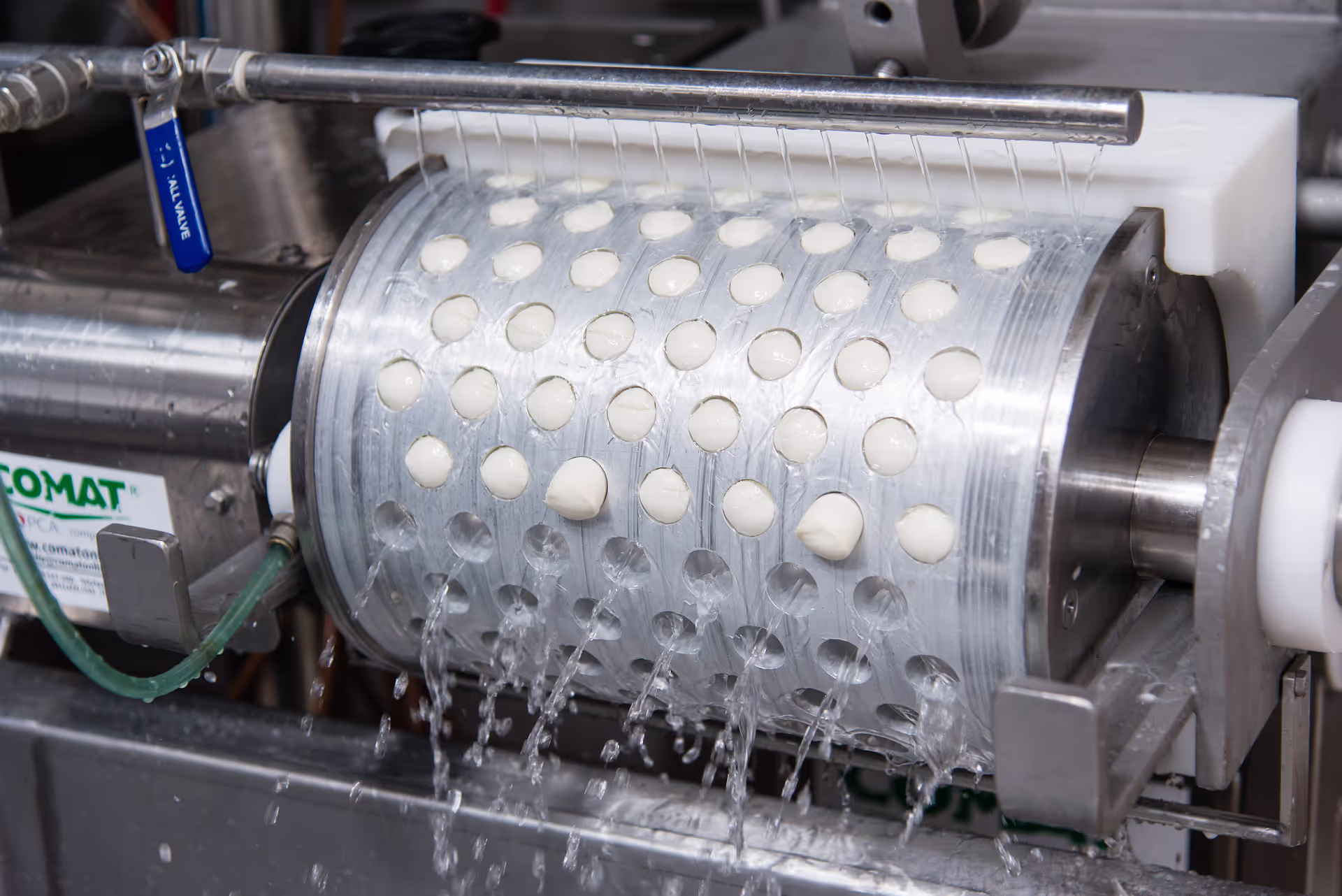 Close-up of a mozzarella cheese production machine. A rotating stainless steel drum with perforations shapes small mozzarella curds while water streams rinse them during the cheese-making process.