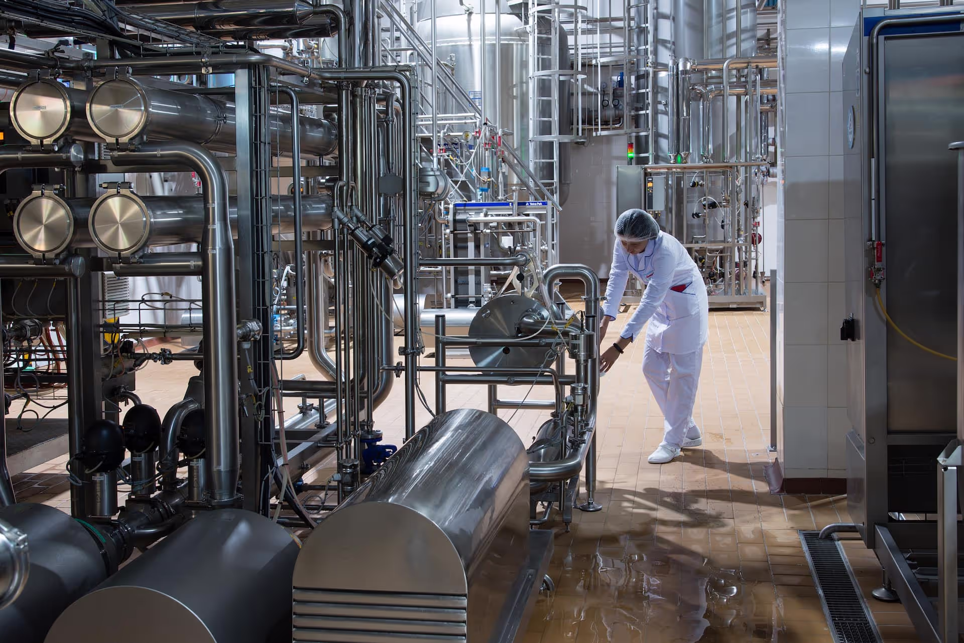 A worker in a white lab coat, hairnet, and gloves inspecting stainless steel machinery in a modern dairy processing facility. The facility is equipped with interconnected pipes, tanks, and control systems.