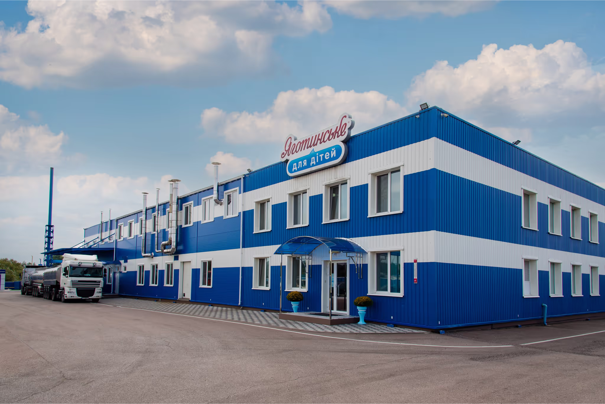 Exterior view of the ‘Яготинське для дітей’ (Yahotynske for Children) dairy production facility. The building is painted in blue and white stripes, with a company sign on top. A milk tanker truck is parked near the entrance, and ventilation pipes are visible along the side of the factory.