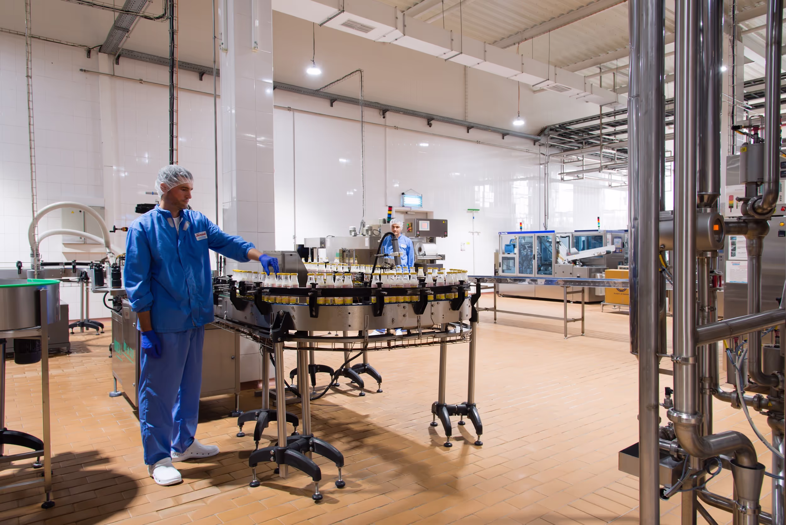 Workers in blue protective clothing, gloves, and hairnets overseeing a dairy production line in a modern factory. Bottles of dairy products with yellow lids move along a conveyor system, surrounded by stainless steel machinery and automated packaging equipment.