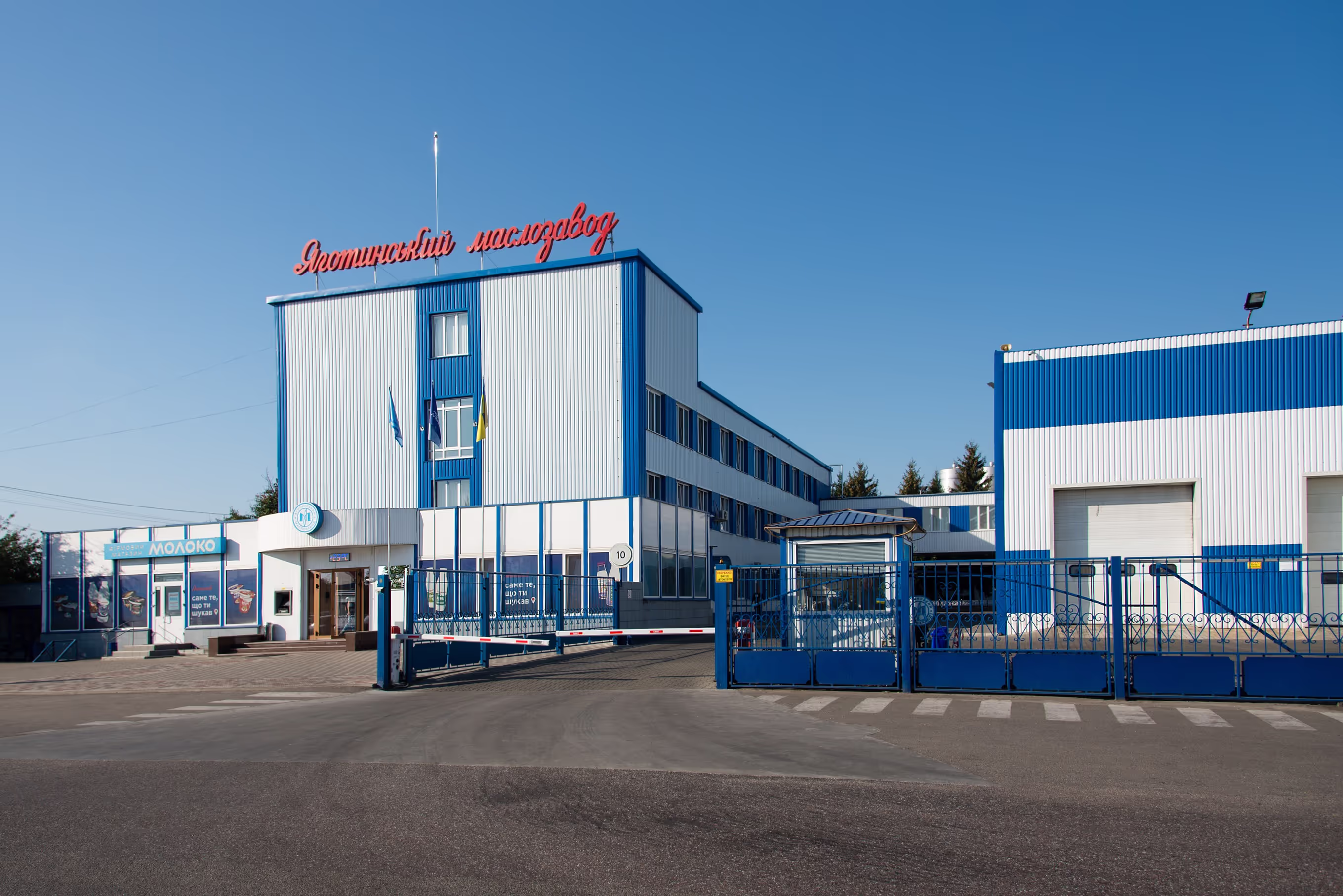 Exterior view of the ‘Яготинський маслозавод’ (Yahotynskyi Butter Plant), a dairy production facility in Ukraine. The blue and white industrial building has a red sign on top, a security gate, and a small store labeled ‘Молоко’ (Milk) at the entrance.
