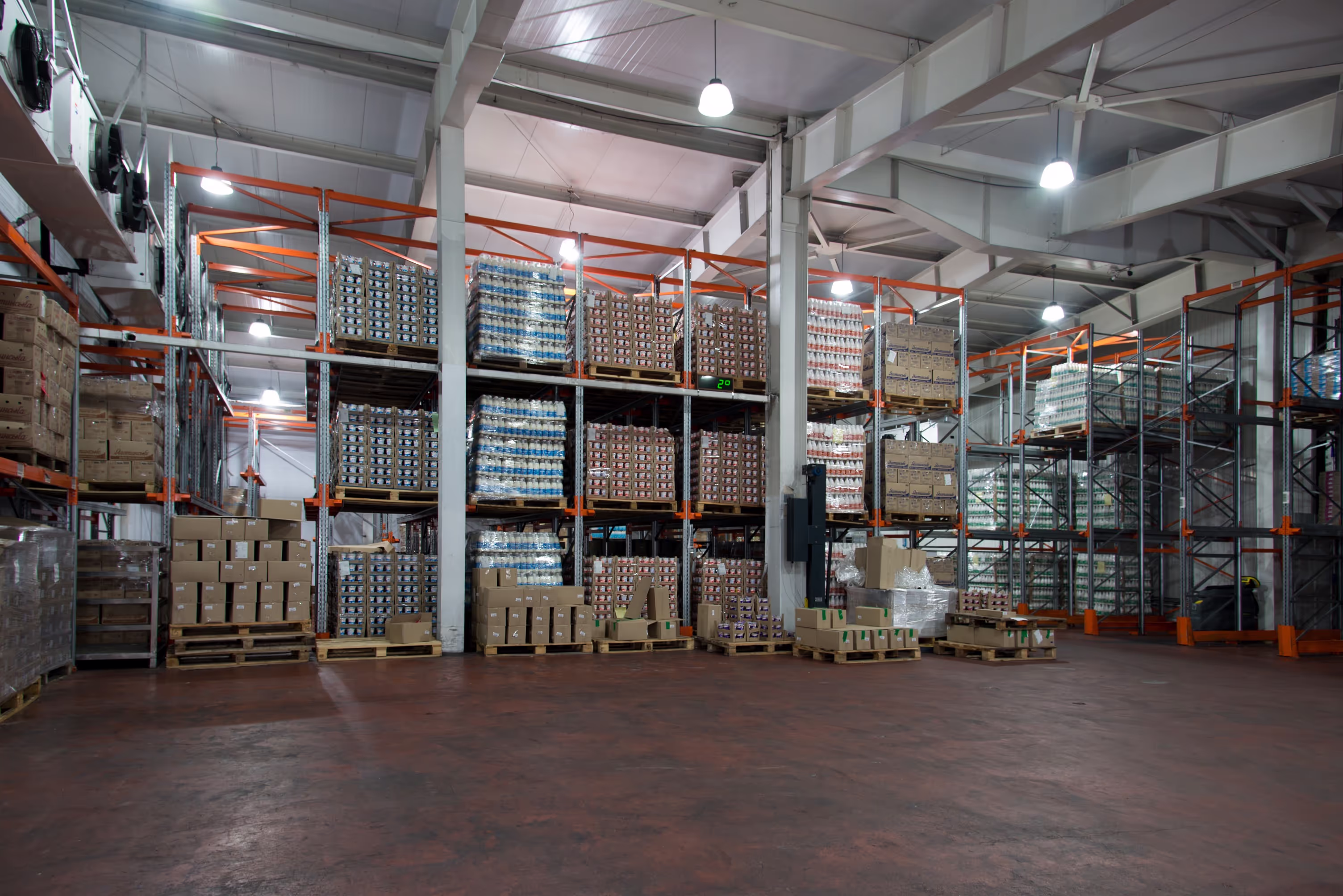 Large warehouse with tall metal racks storing dairy products in bulk. The shelves are stacked with pallets of packaged goods, including yogurt and milk, wrapped in plastic. Cardboard boxes and wooden pallets are organized on the floor, with a temperature display showing ‘2°C’ for cold storage.