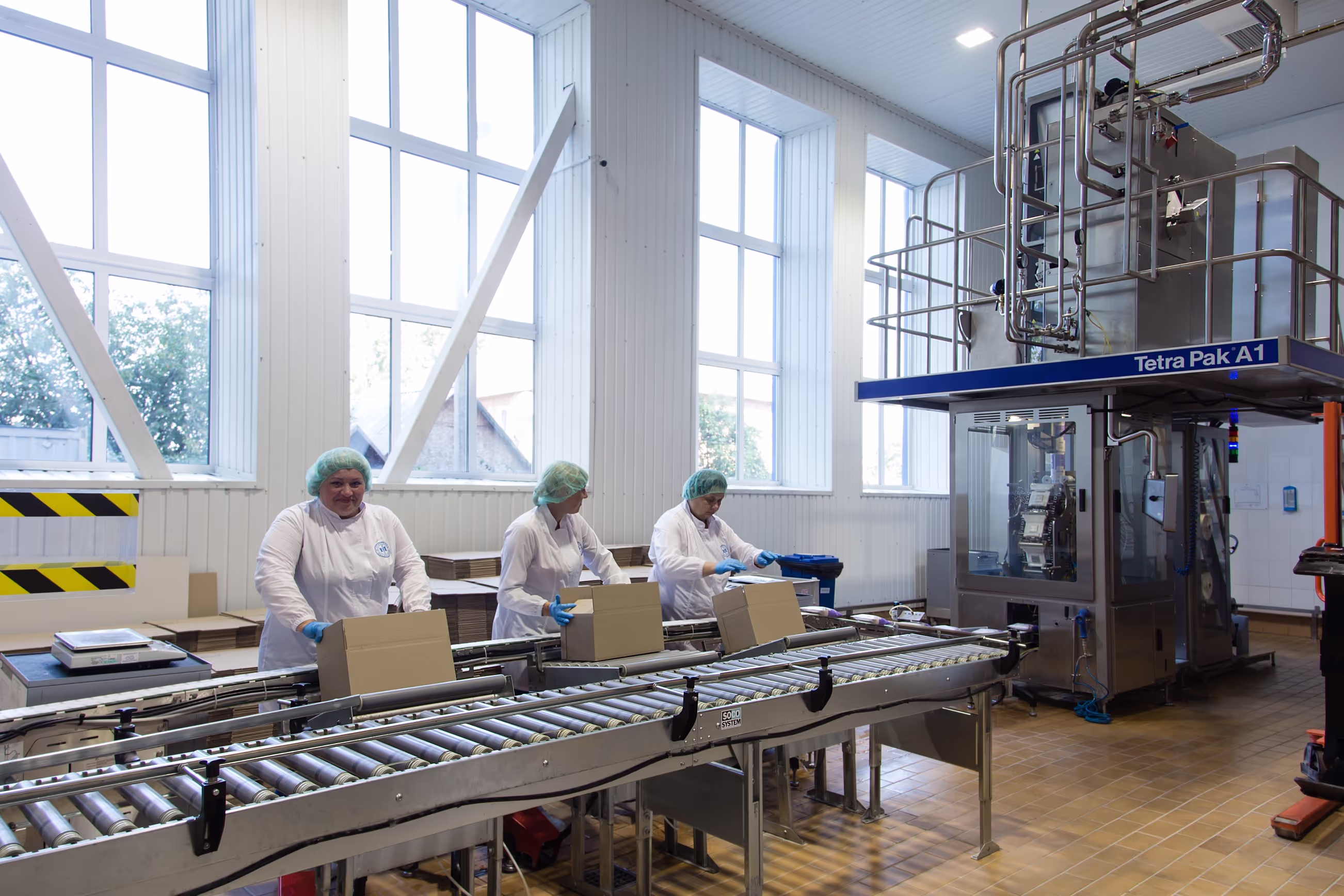 Workers in white uniforms, hairnets, and gloves assembling and sealing cardboard boxes on a conveyor belt in a dairy packaging facility. A large Tetra Pak A1 machine is visible on the right, used for automated packaging of dairy products.