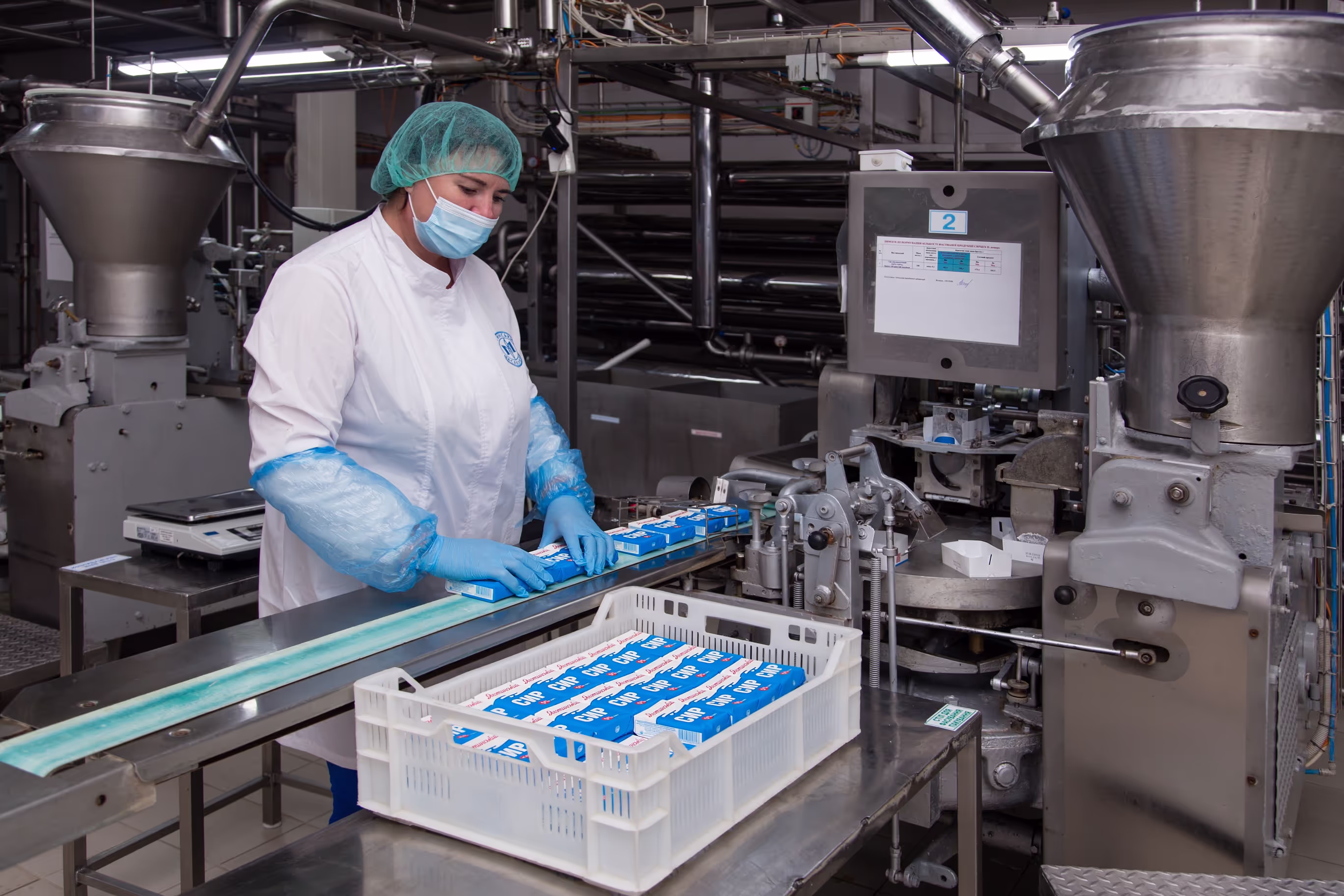 A worker in a white uniform, blue gloves, and protective sleeves handling packaged butter on a conveyor belt in a dairy production facility. The butter packages are labeled ‘Сир’ (Cheese) and are being placed into a plastic crate for further processing.