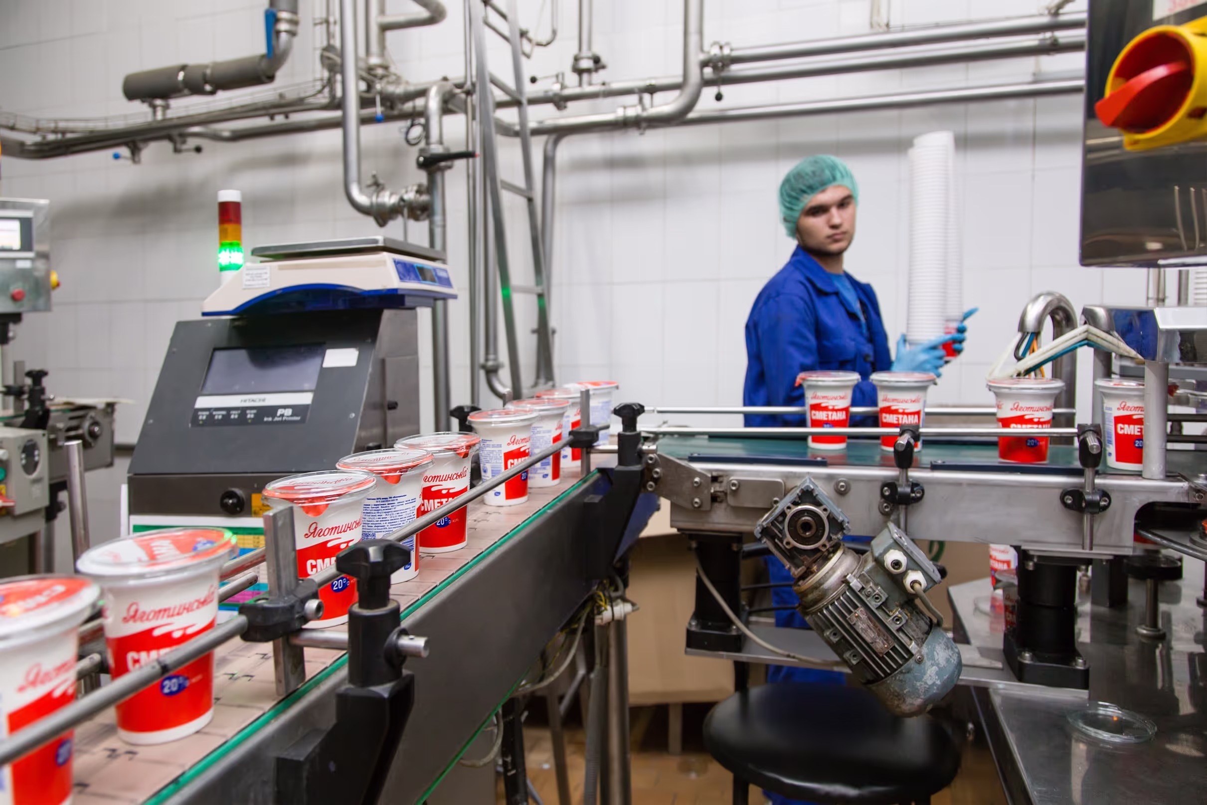 A worker in a blue uniform and hairnet monitoring the automated production line of ‘Яготинська Сметана’ (Yahotynska Sour Cream) in a dairy processing facility. Plastic cups of sour cream move along a conveyor belt, passing through a quality control and packaging station.