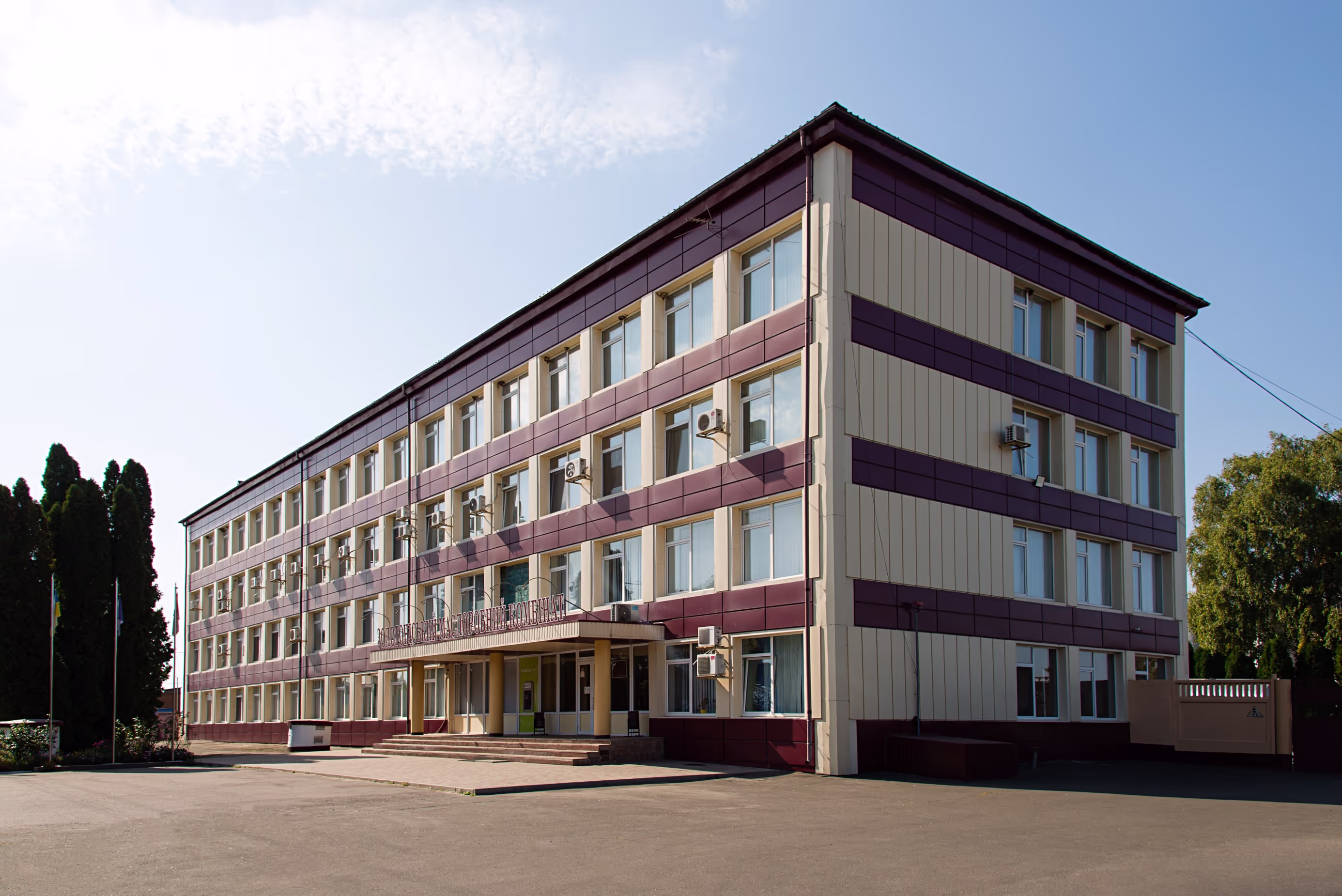 Exterior view of the administrative building of ‘Золотоніський маслоторговий комбінат’ (Zolotonosha Butter Trading Plant). The multi-story structure features beige and maroon panels, multiple windows with air conditioning units, and a main entrance with a canopy. Trees and flags are positioned in front of the building.