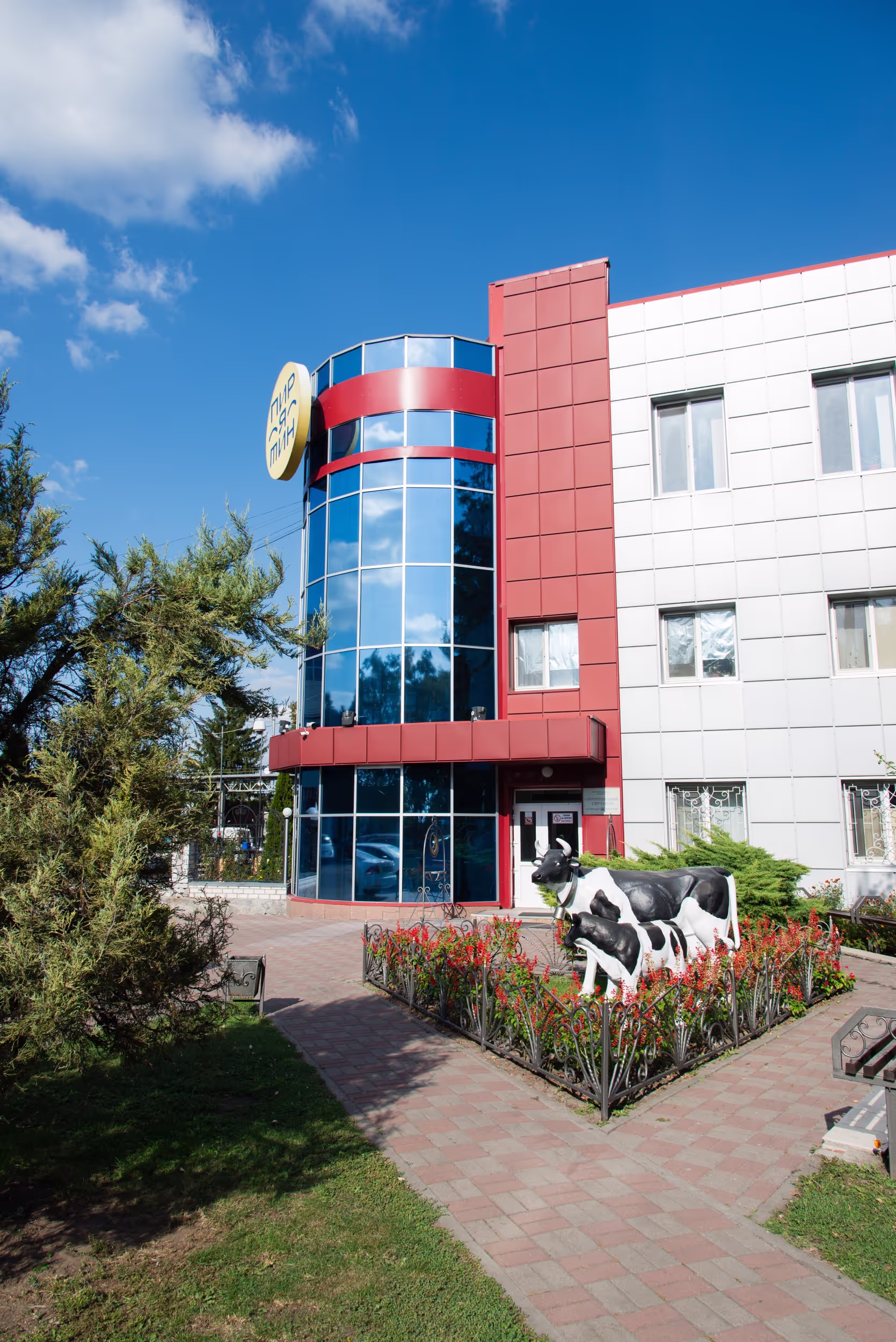 Entrance to a dairy facility with a landscaped area featuring life-sized black and white cow statues surrounded by red flowers. The building has large glass windows, a red and white facade, and a sign near the door. Trees and greenery decorate the surroundings.