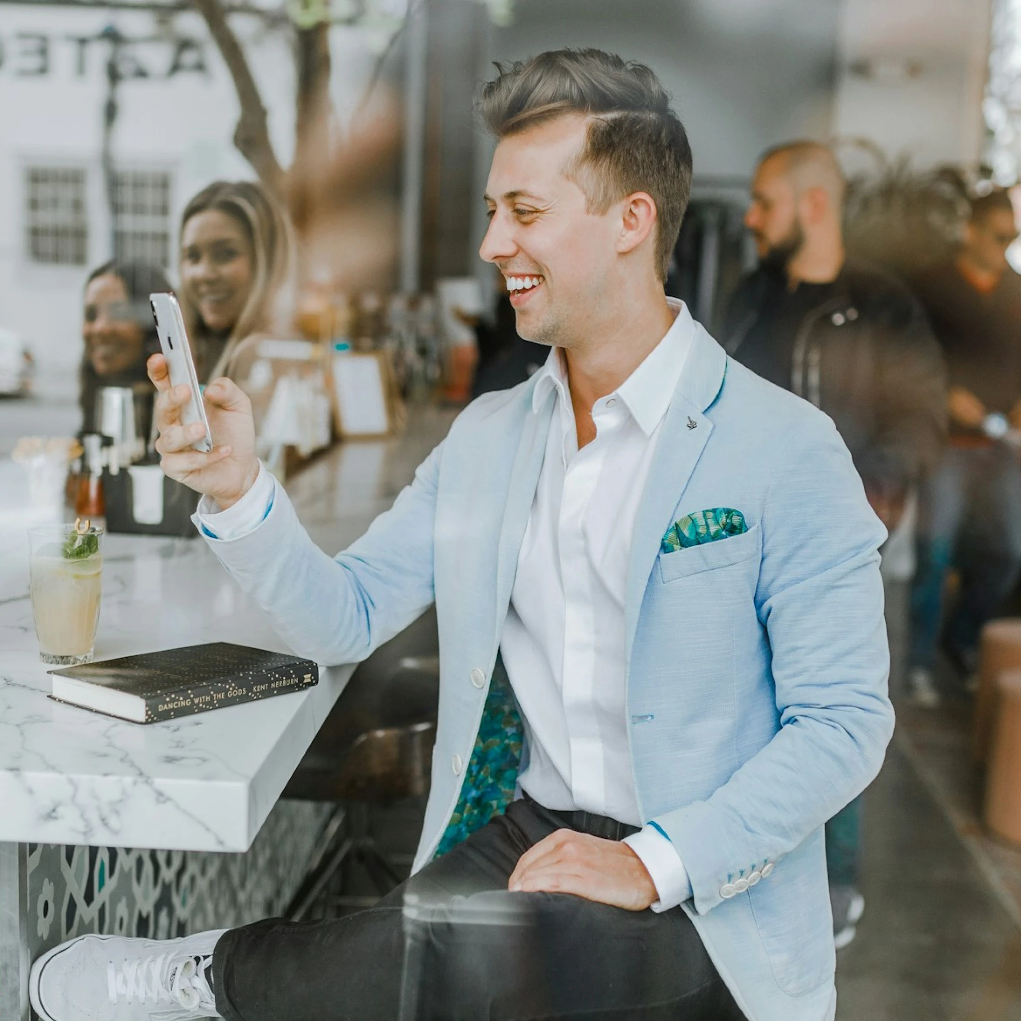 A stylish man in a blue blazer, smiling and taking a selfie with his smartphone at a café, with other people visible behind him.