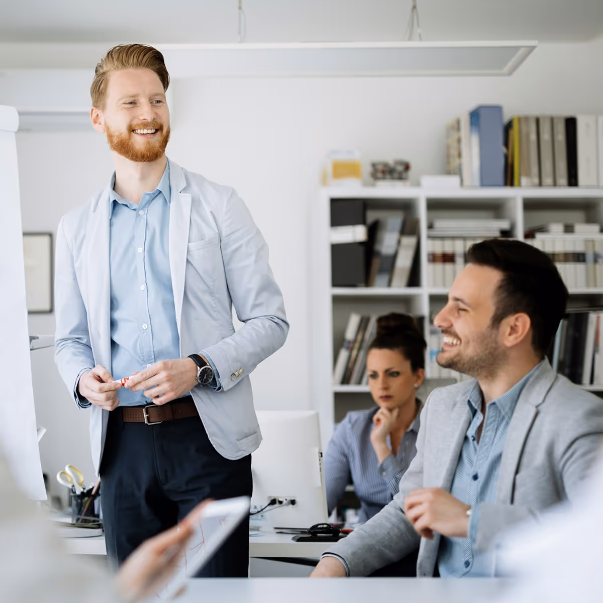 Three colleagues in a modern office, smiling and working near a flip chart and computer—capturing the energy of shared ideas and takeaway resources.