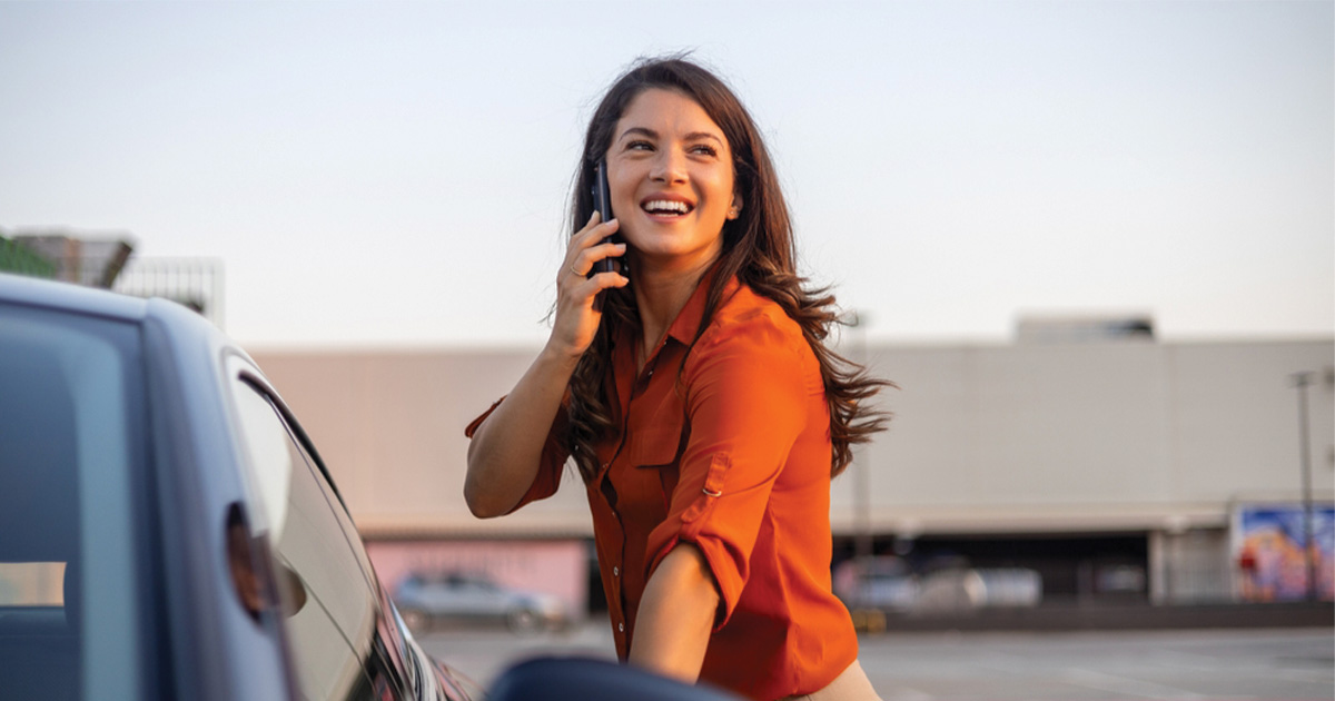 Smiling woman in orange shirt talking on phone next to car at dealership – Carmada customer experience