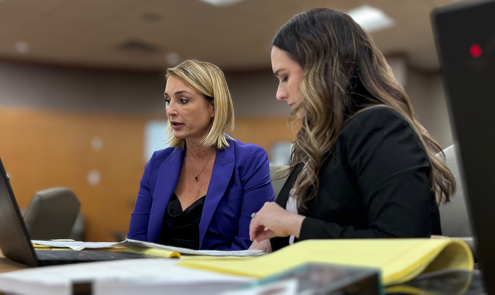 Two professional women in business attire working intently together reviewing documents and using a laptop in an office setting.
