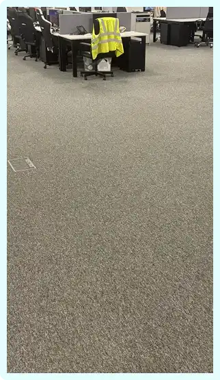 Office workspace with carpeted floor, rows of desks, office chairs, and a high-visibility safety vest draped over one chair.