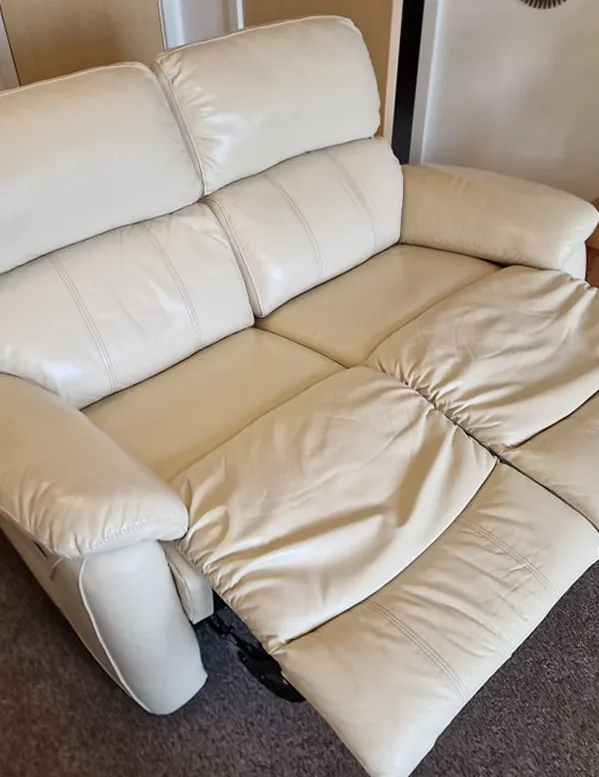 Cream-colored leather recliner sofa with one footrest extended on a carpeted floor indoors.