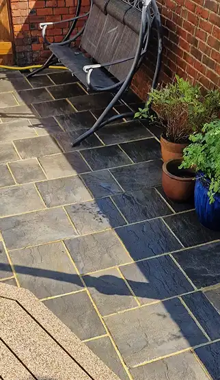 Outdoor patio with clean slate tiles, a black metal swing, and potted plants by a brick wall.