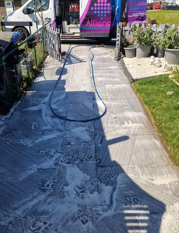 Driveway being professionally cleaned with foam detergent and a hose visible in front of a cleaning van.
