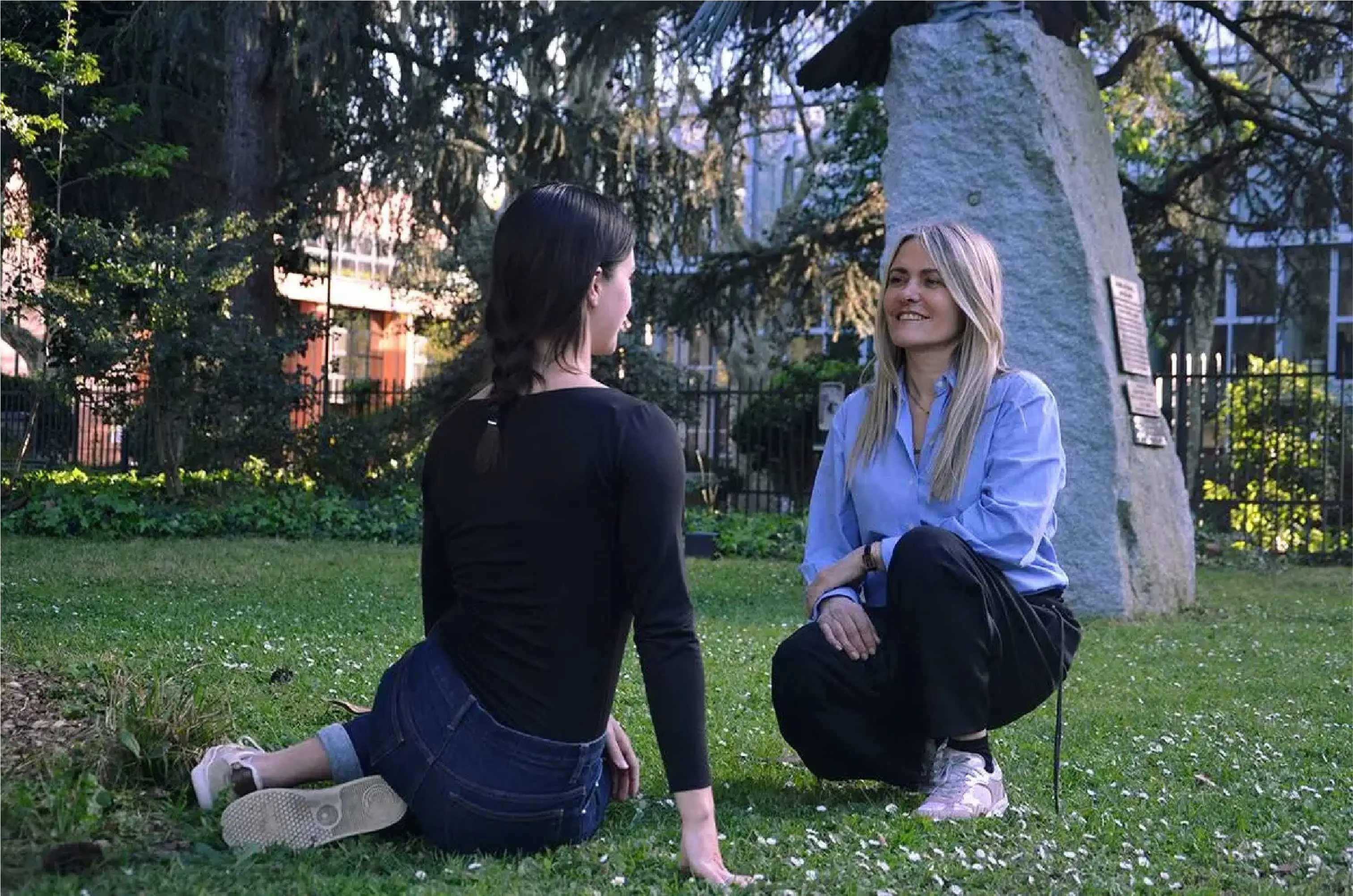 Two women sitting on grass in a park, engaged in a friendly conversation on a sunny day.