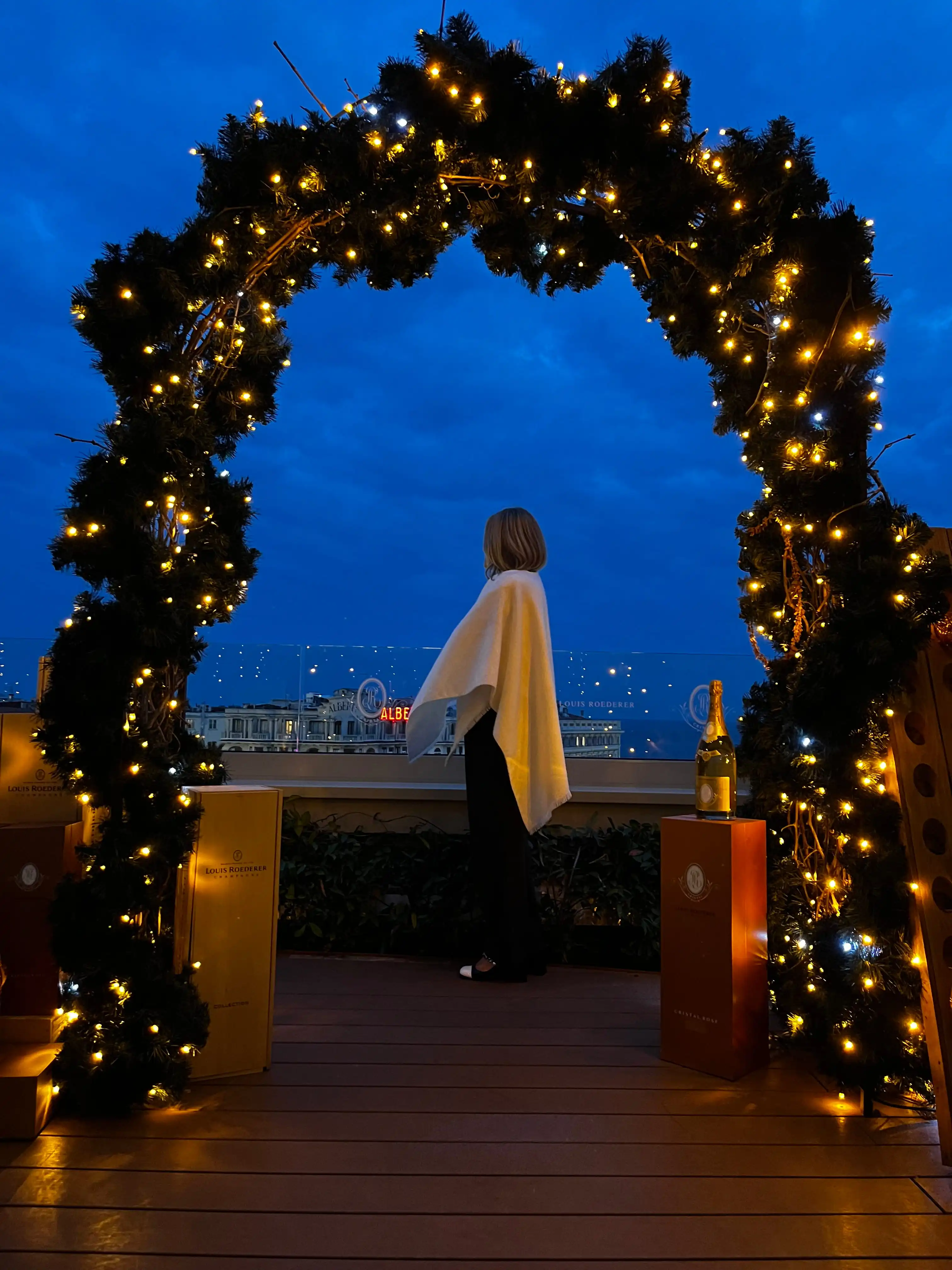 Personne debout sous un arc de guirlandes lumineuses sur une terrasse au crépuscule.