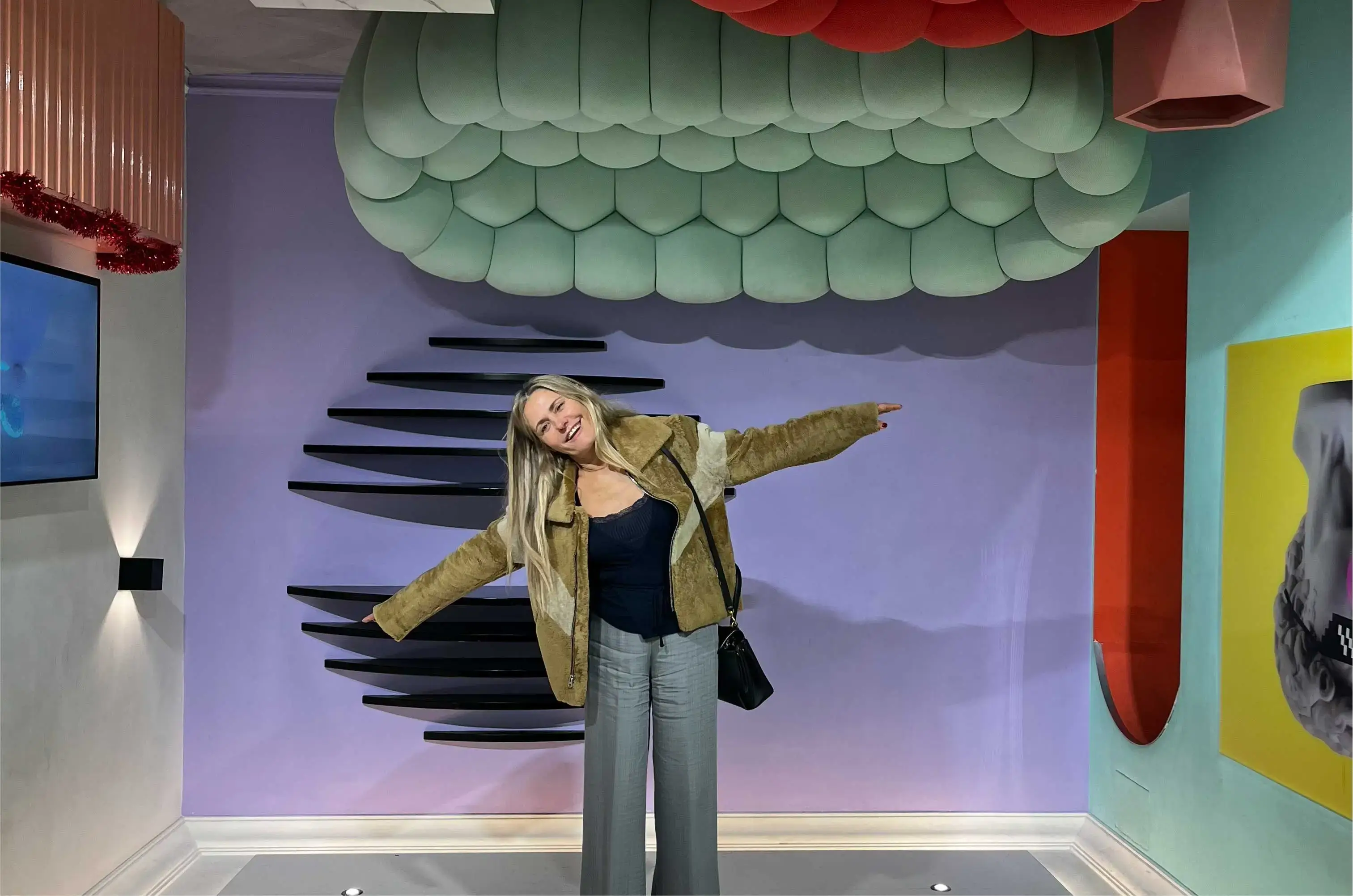 Femme souriante avec veste et pantalon ouvrant les bras devant un mur violet avec décoration moderne.