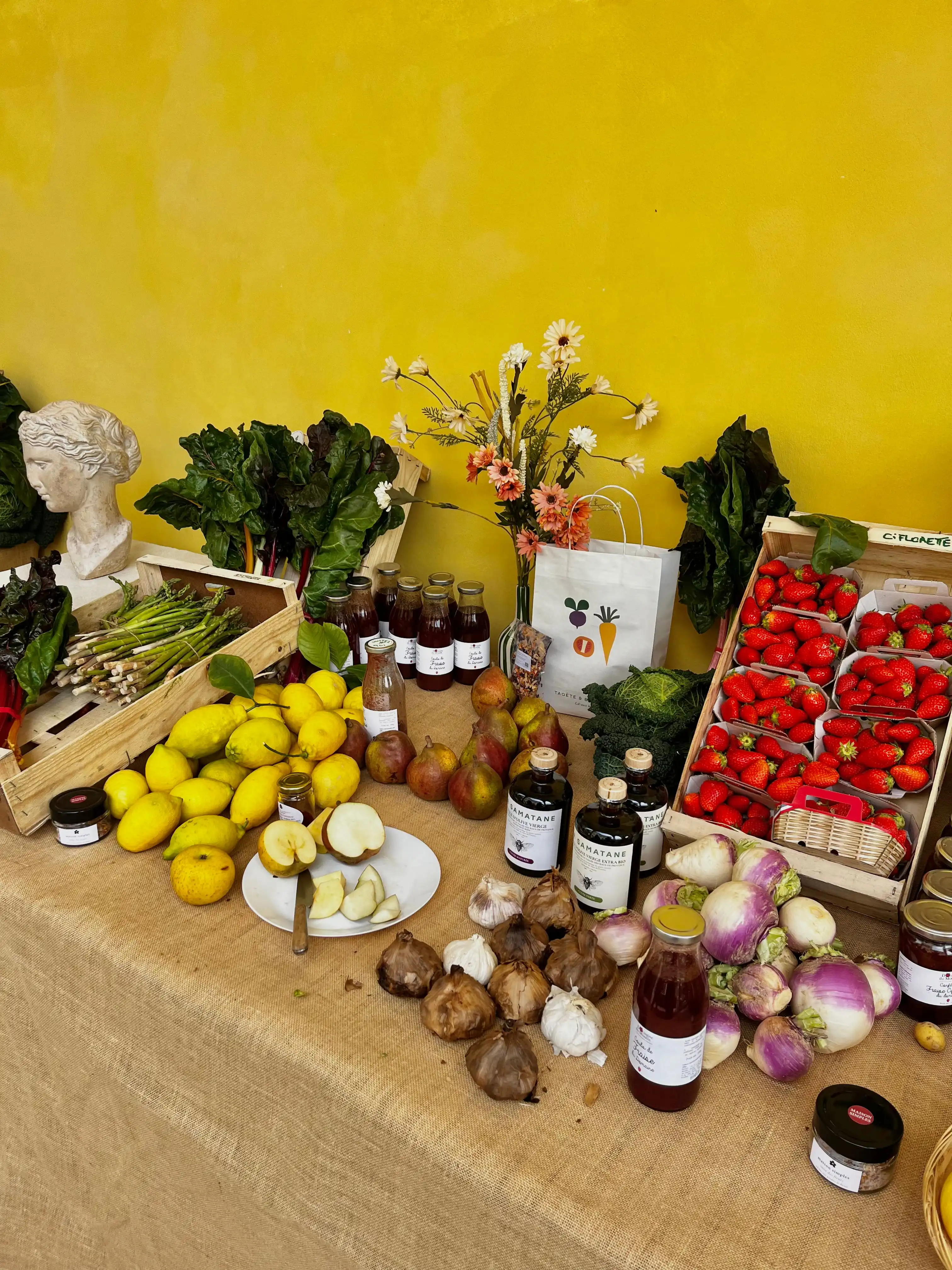 Fruits et légumes frais, jus et fleurs sur une table couverte de toile de jute jaune.