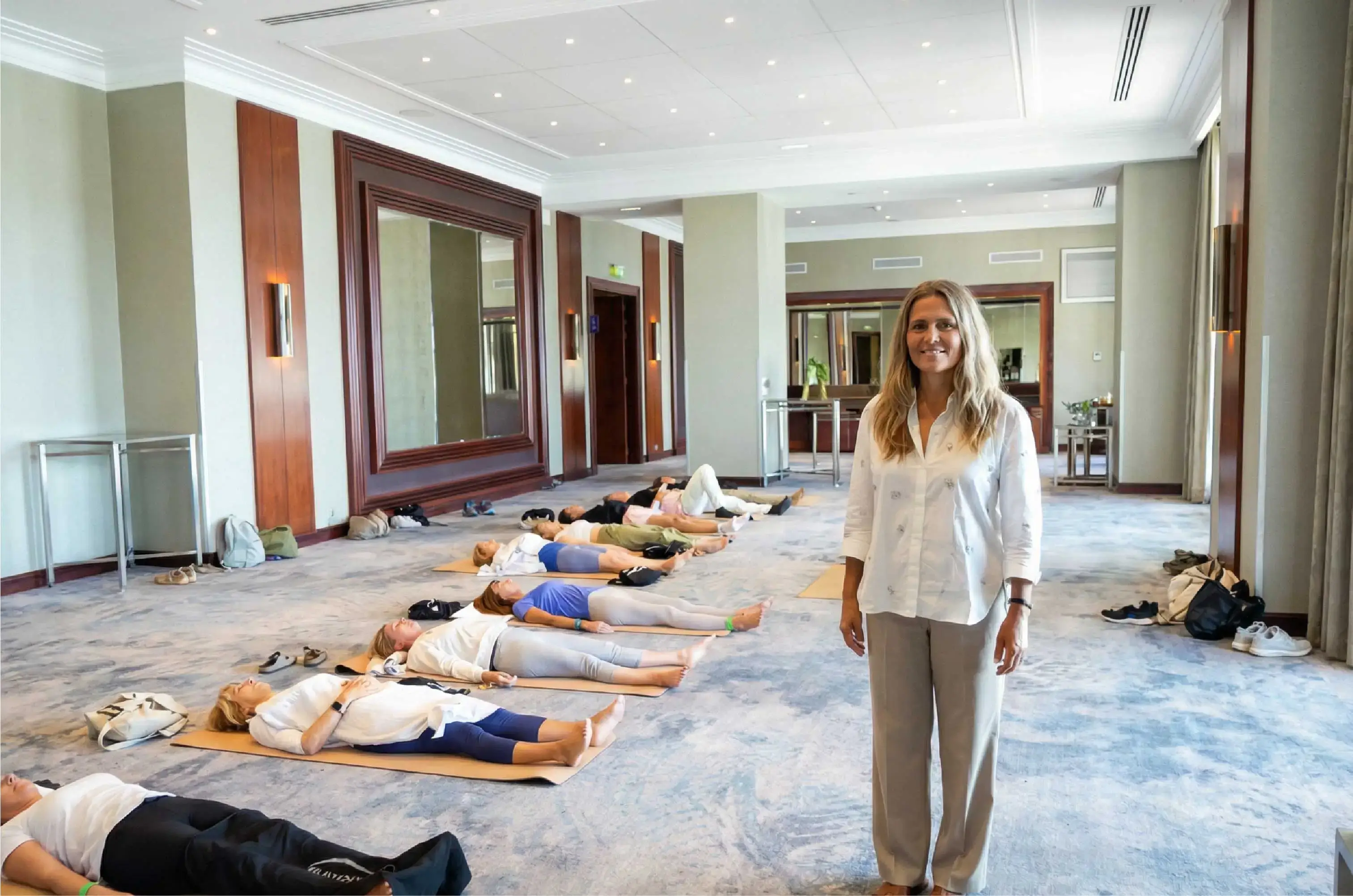 Femme debout souriante dans salle avec personnes allongées sur tapis pour exercice de relaxation.