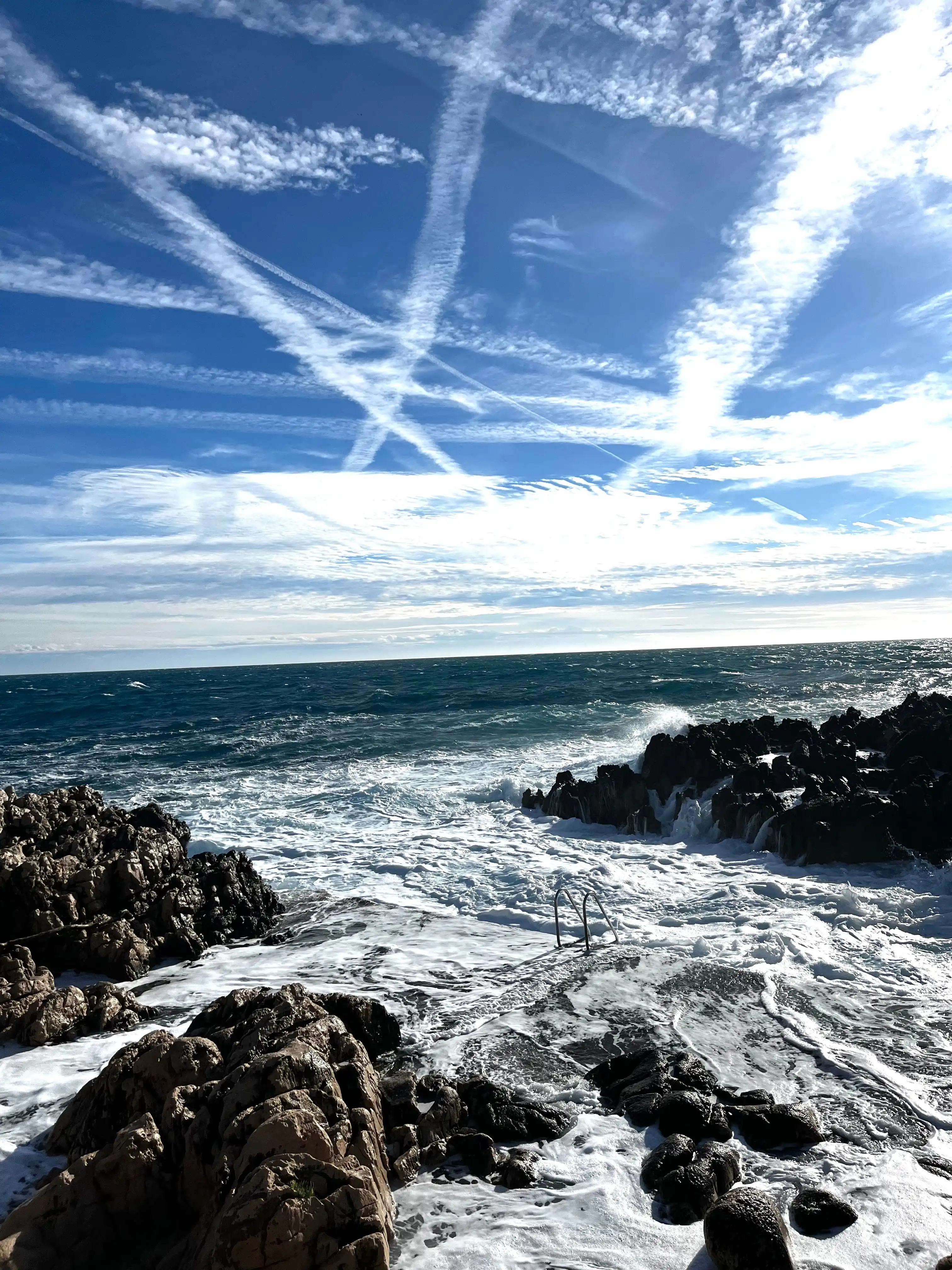 Côte rocheuse avec vagues agitées et ciel bleu traversé de traînées de nuages blancs.