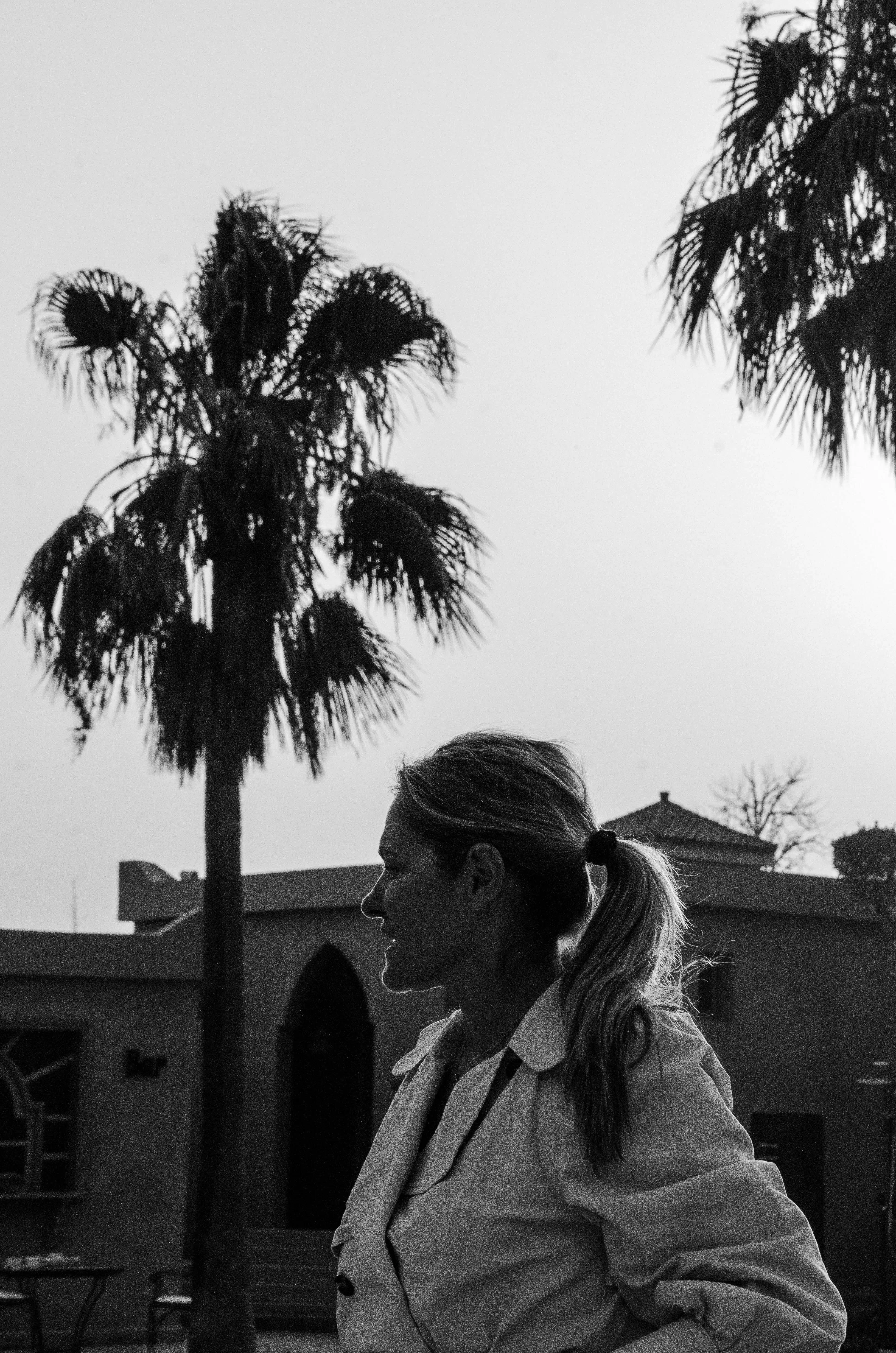 Femme de profil avec une queue de cheval devant des palmiers et un bâtiment au crépuscule.