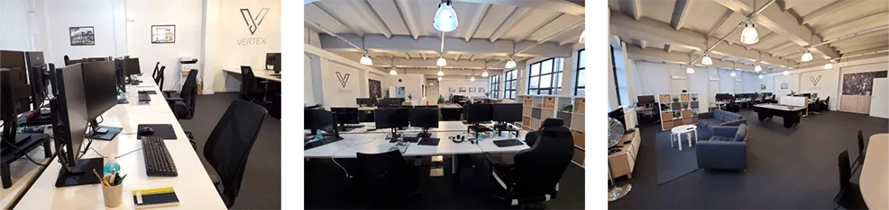 Modern open office space with multiple desks, computer monitors, ergonomic chairs, a lounge area with couches, and vaulted ceilings with exposed beams and hanging lights.