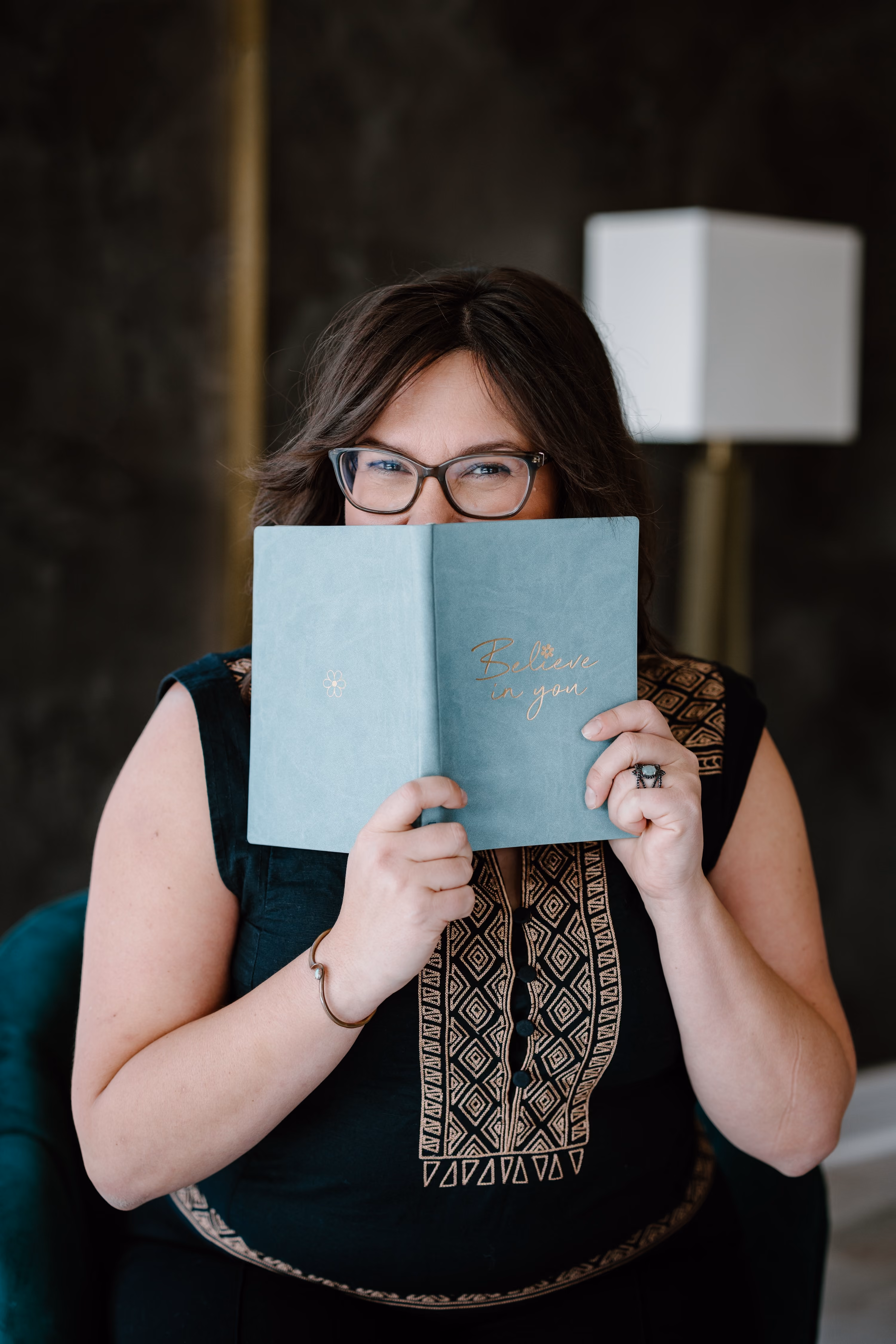 Smiling woman with glasses holding a closed light blue journal with the words 'Believe in you' written in gold lettering, partially covering her face, seated in a room with a dark background and a white table lamp.