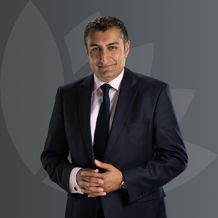 A man in a dark suit and tie standing with his hands clasped, against a grey background