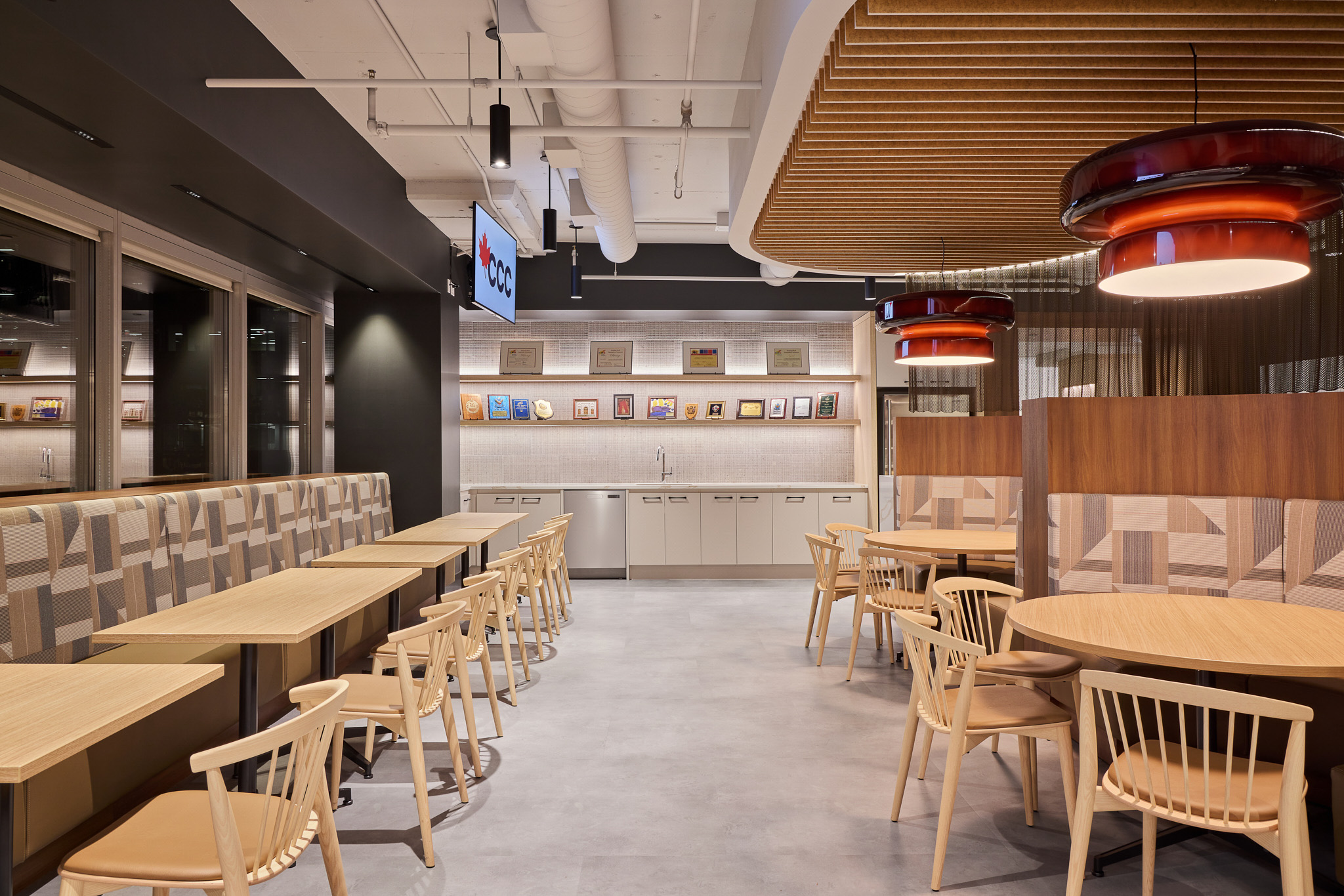 Interior office dining space featuring booths, open chair seating with a kitchenette set up along the back, with neutral tones and wood finishes.