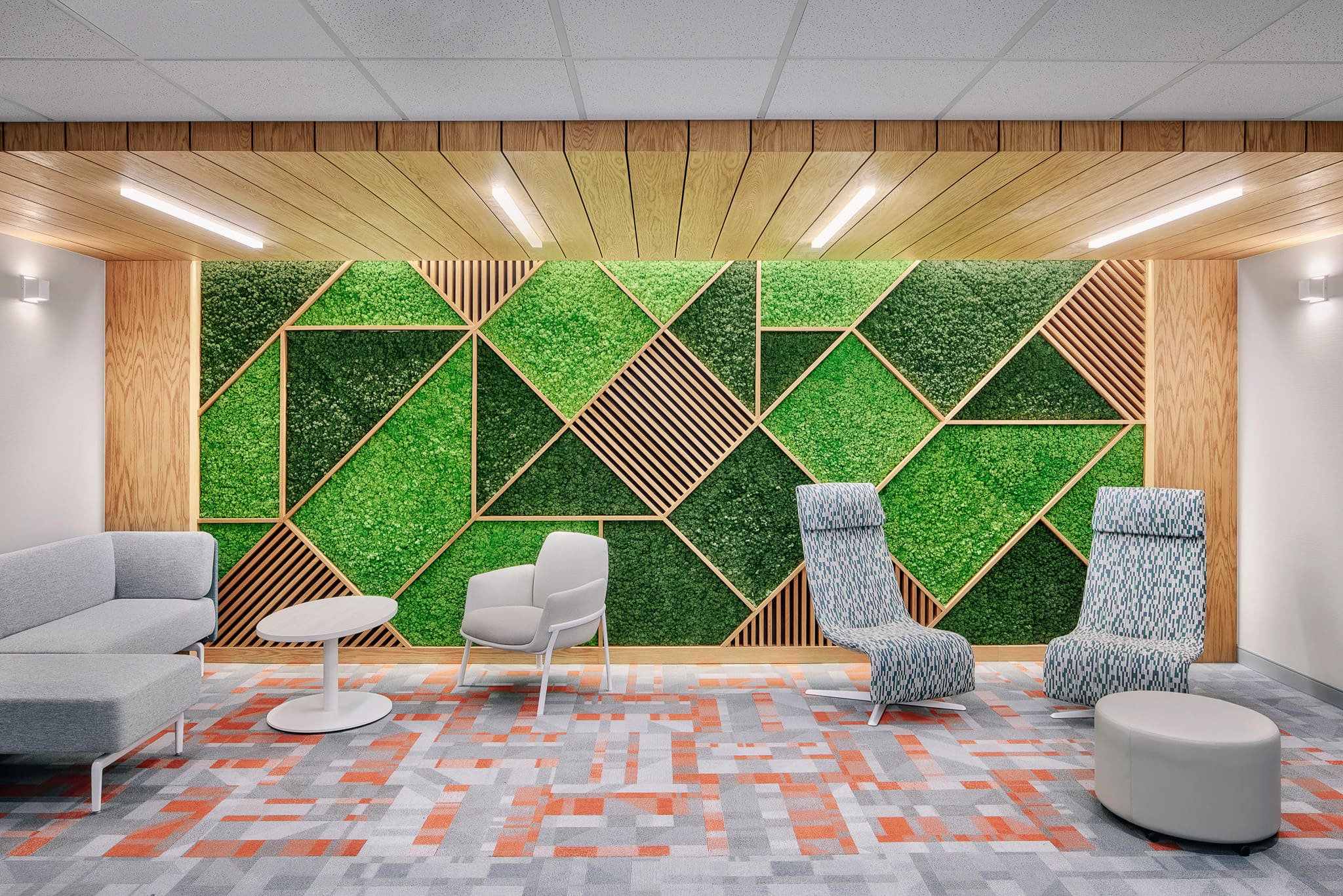 Modern interior office space seating area featuring a sofa, table, chairs, and an accent wall with wood and green plants in a geometric pattern