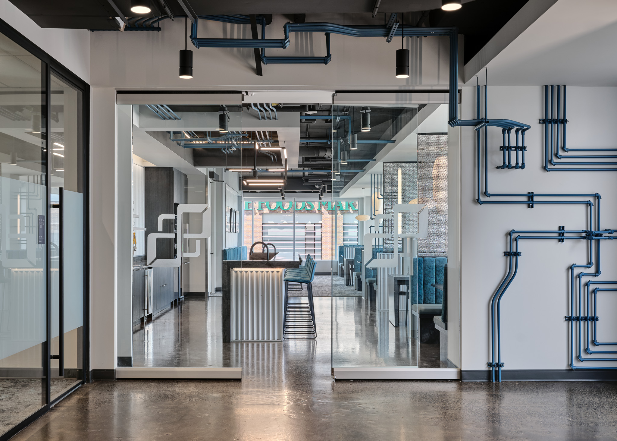 Modern interior office space featuring glass doors, a unique visible pipe formation along the wall, monochrome tone with whites, grays, and blacks with a blue accent. 