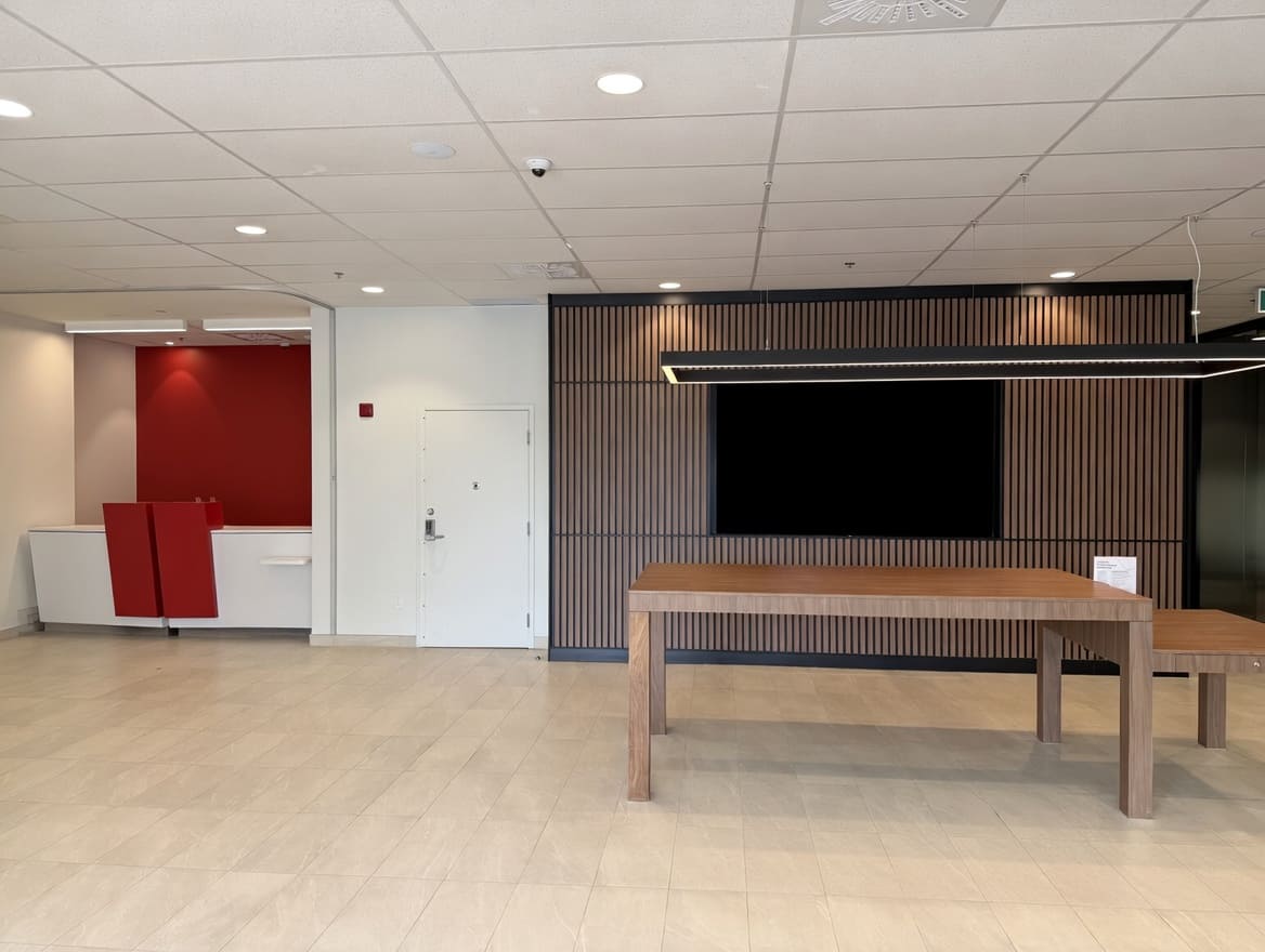 Open concept office space with a desk on the left-hand side, a door in the middle, and a meeting space featuring a TV, table, and minimalistic rectangle lighting. 