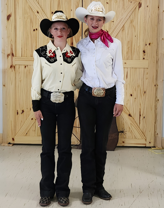 Two women dressed as rodeo royalty in western attire with hats, decorative shirts, and large belt buckles, standing indoors in front of wooden doors.