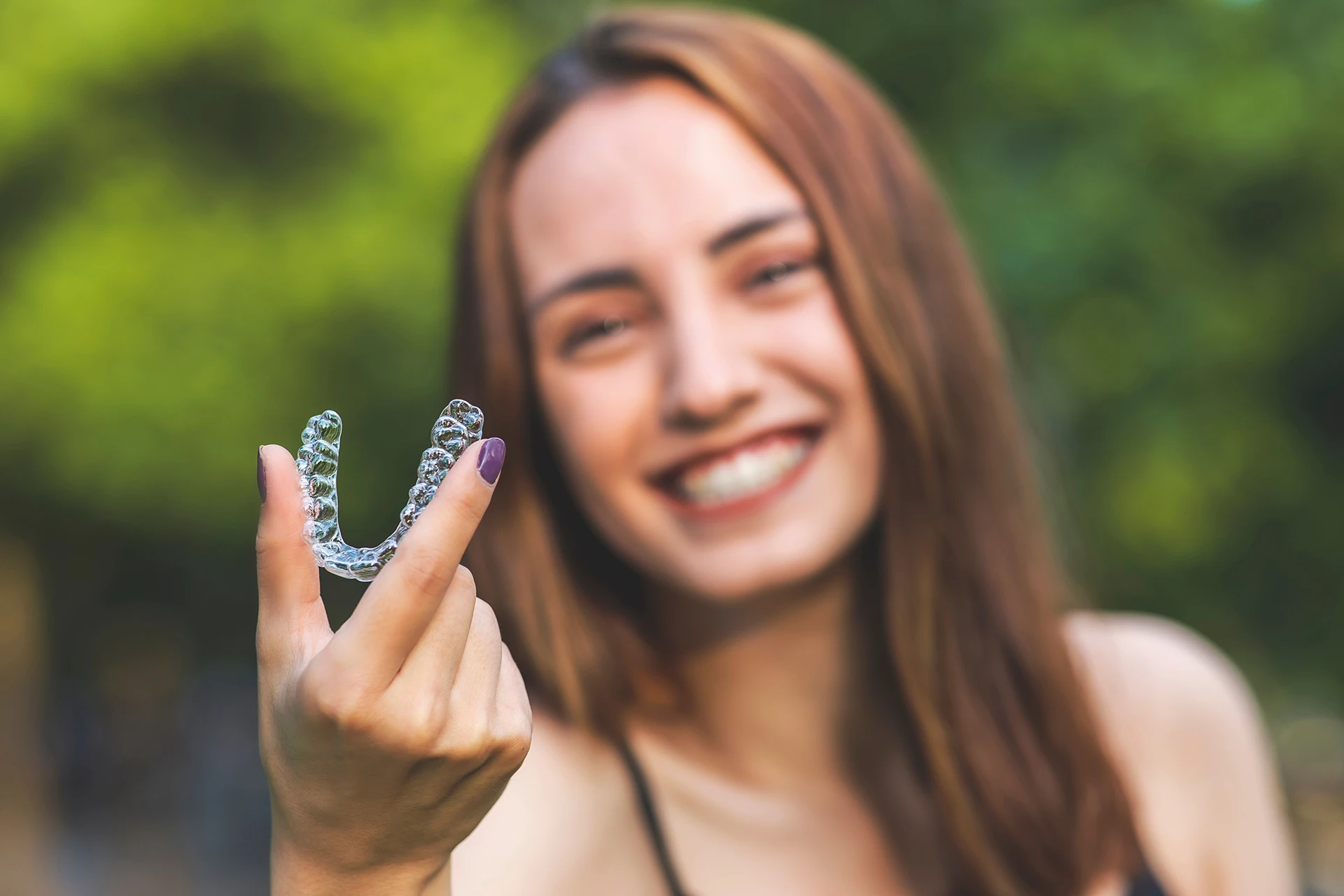 Smiling woman holding a clear dental aligner outdoors with blurred green background.