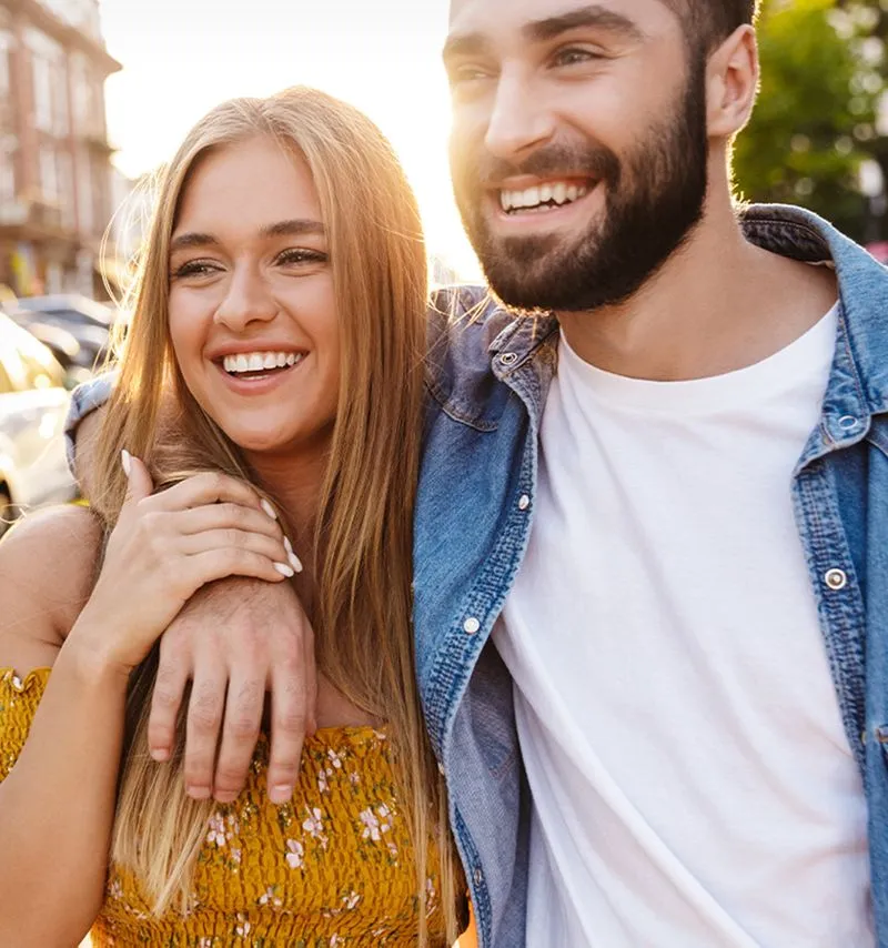Smiling young couple outdoors with the man’s arm around the woman’s shoulder on a sunny day.