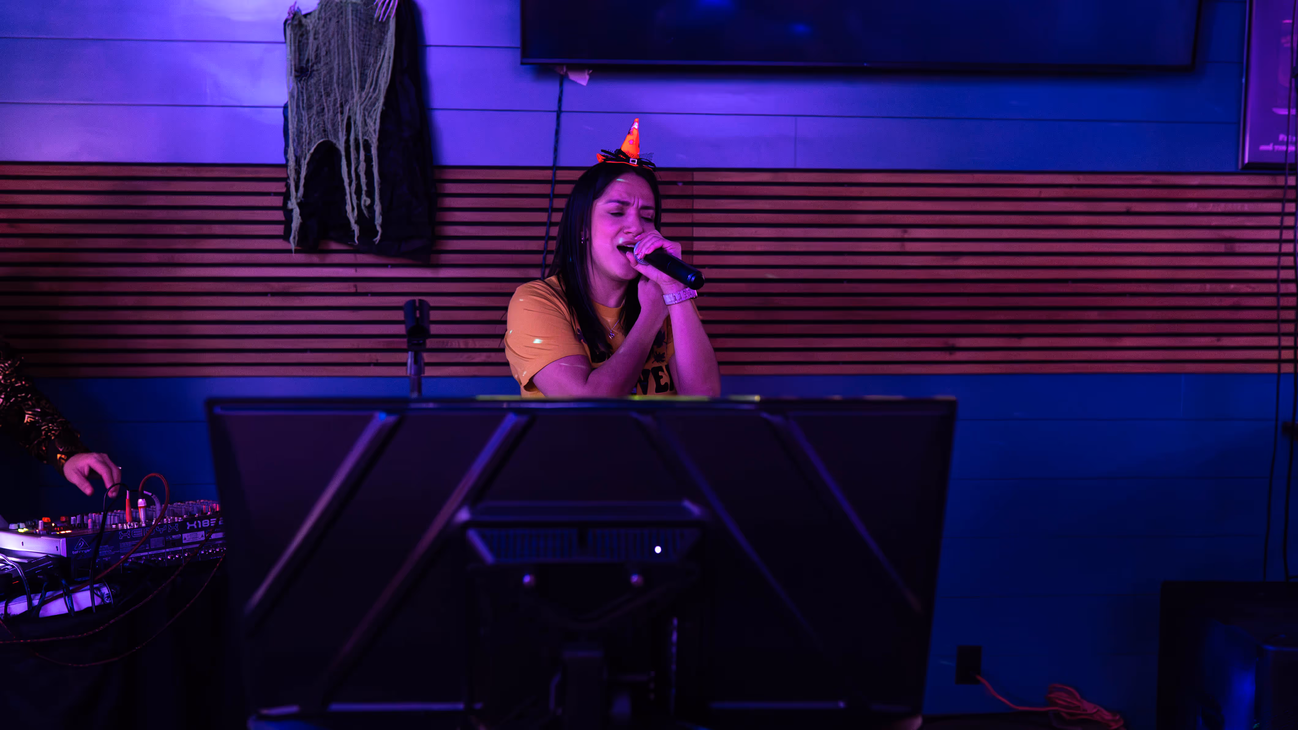 Woman wearing a party hat singing into a microphone in a dimly lit room with purple lighting.