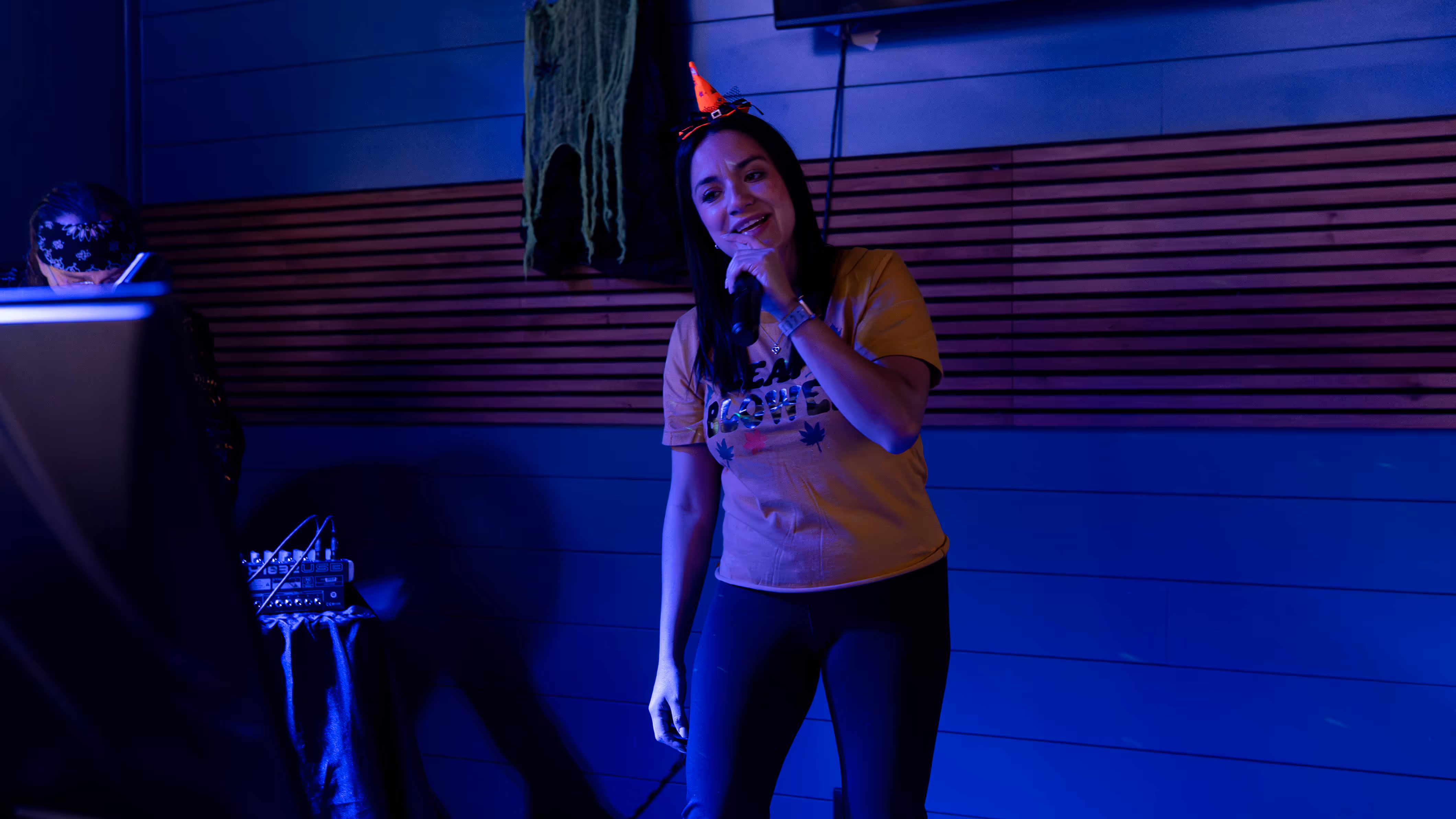 Woman wearing a small orange witch hat and yellow shirt singing into a microphone in a dimly lit room with blue and purple lighting.