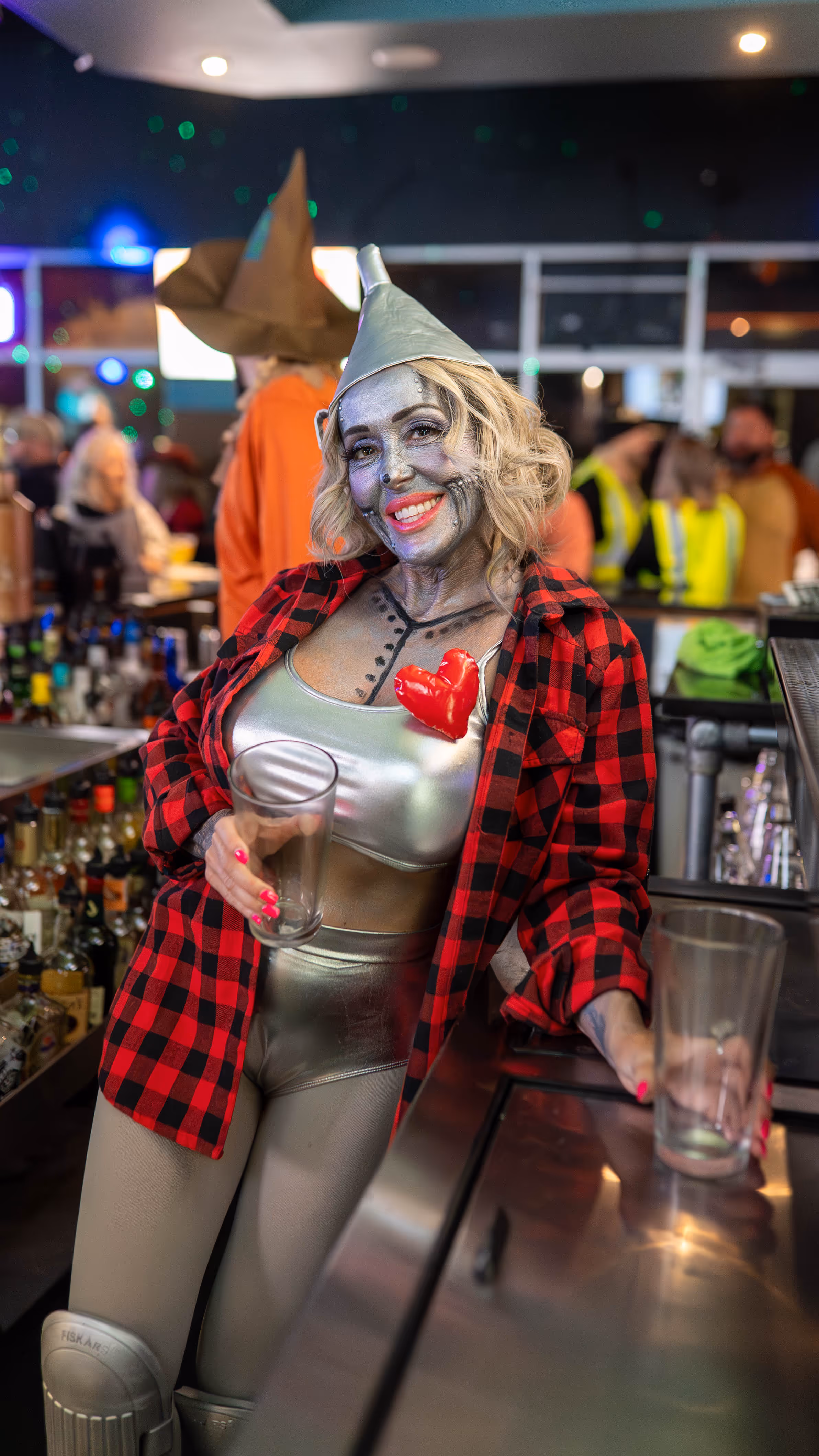 Woman dressed as the Tin Man from The Wizard of Oz with silver face paint, metallic outfit, red plaid shirt, and red heart, holding two empty glasses in a bar.