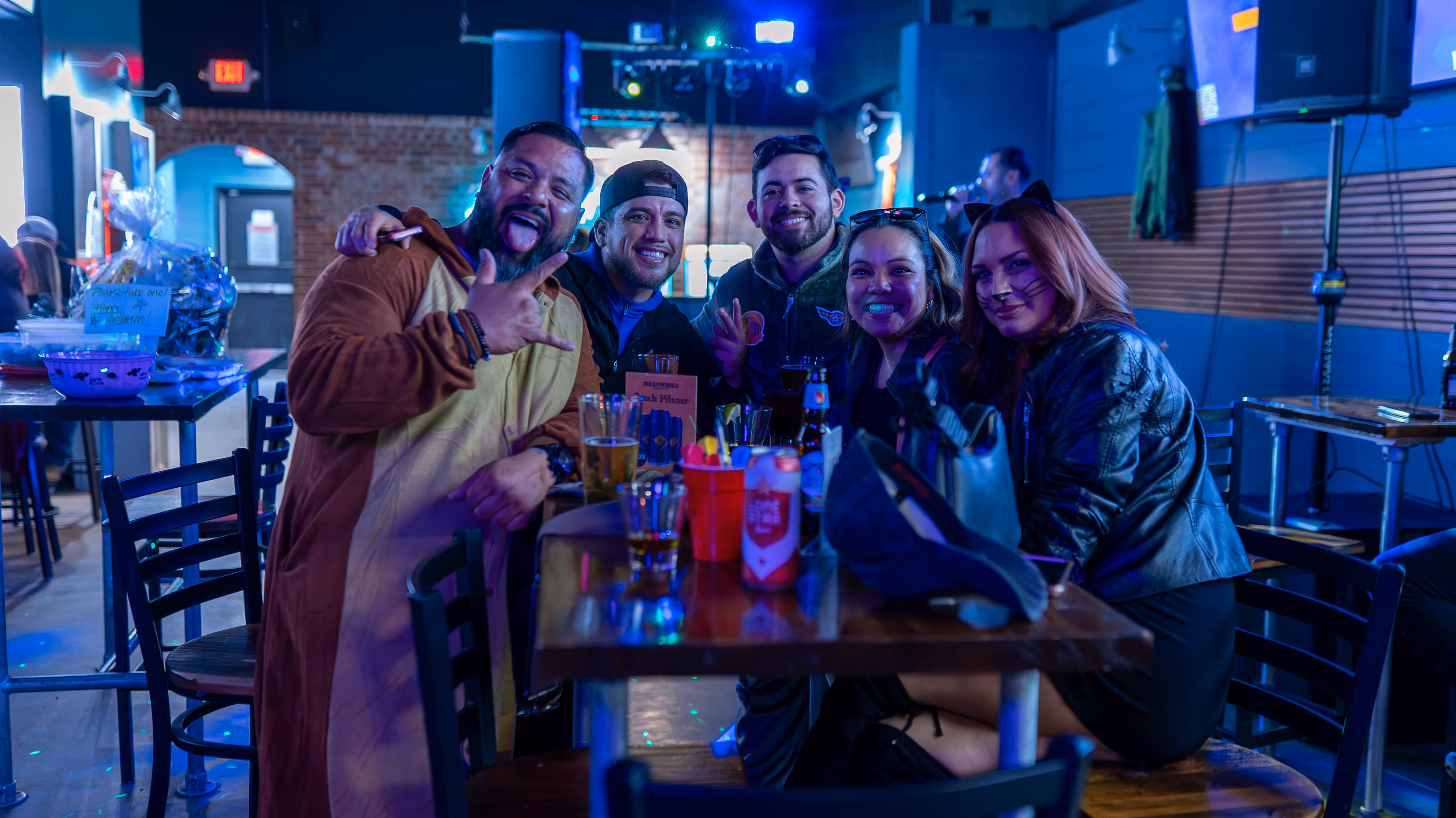 Group of five friends at a bar table smiling, with one person wearing a brown animal costume and another with cat face paint.