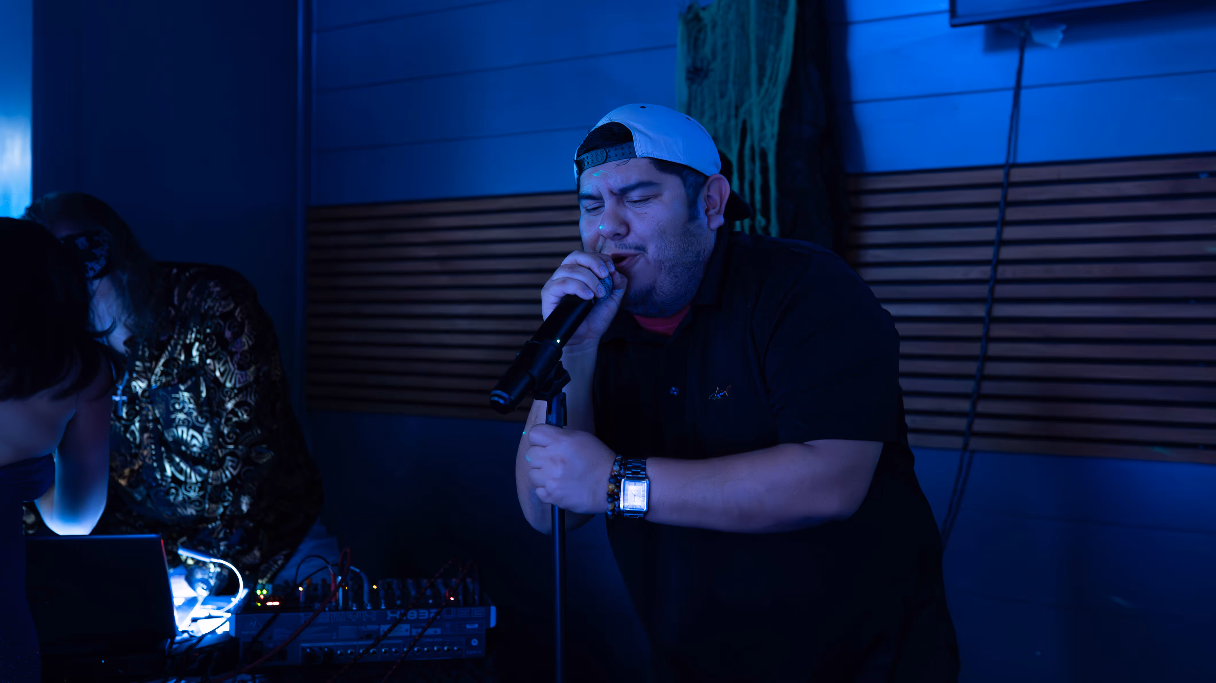 Man wearing a backward white cap singing into a microphone in a dimly lit room with blue lighting.
