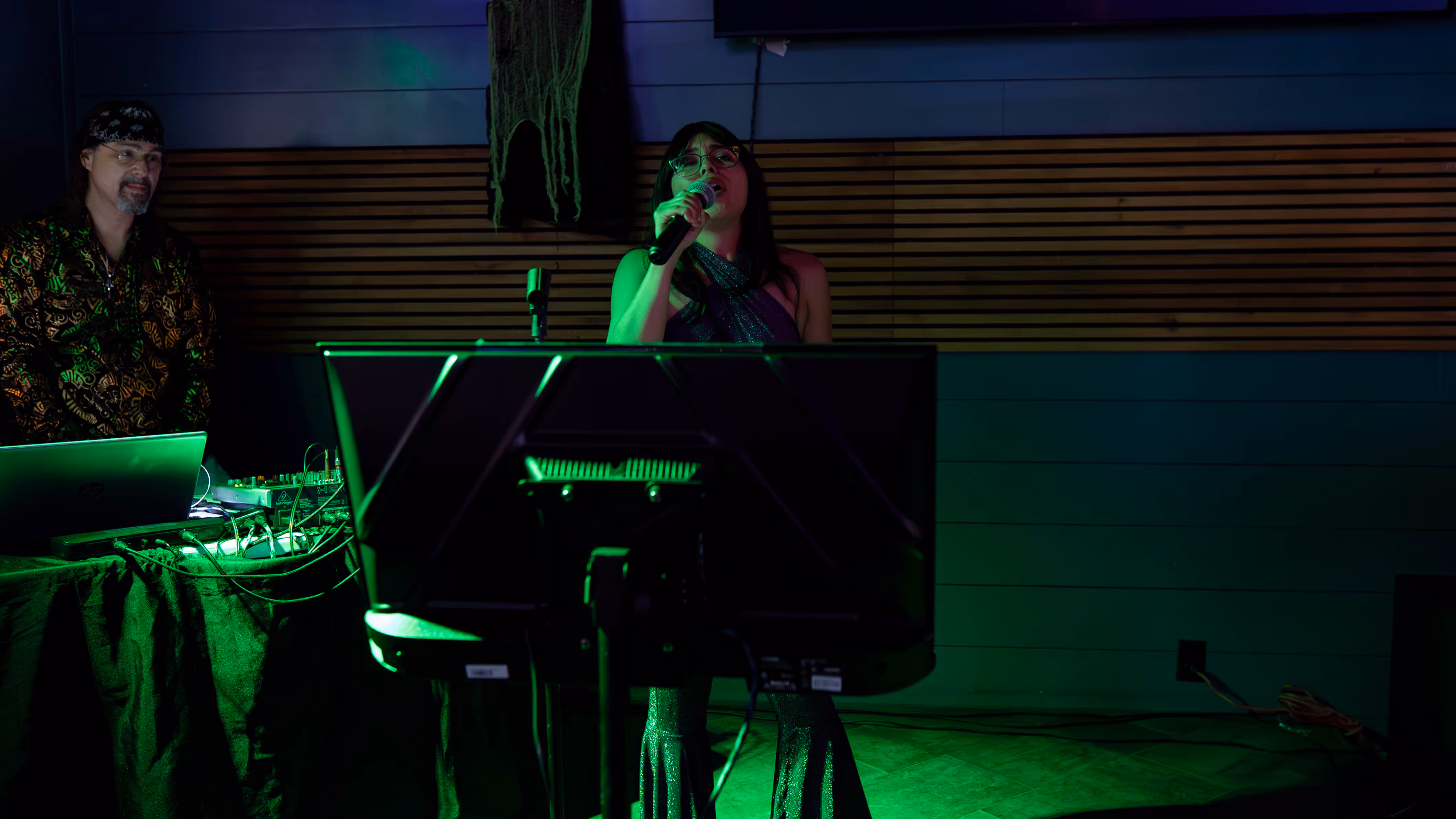 Woman with glasses singing into a microphone in front of a large screen while a man with a bandana operates sound equipment.