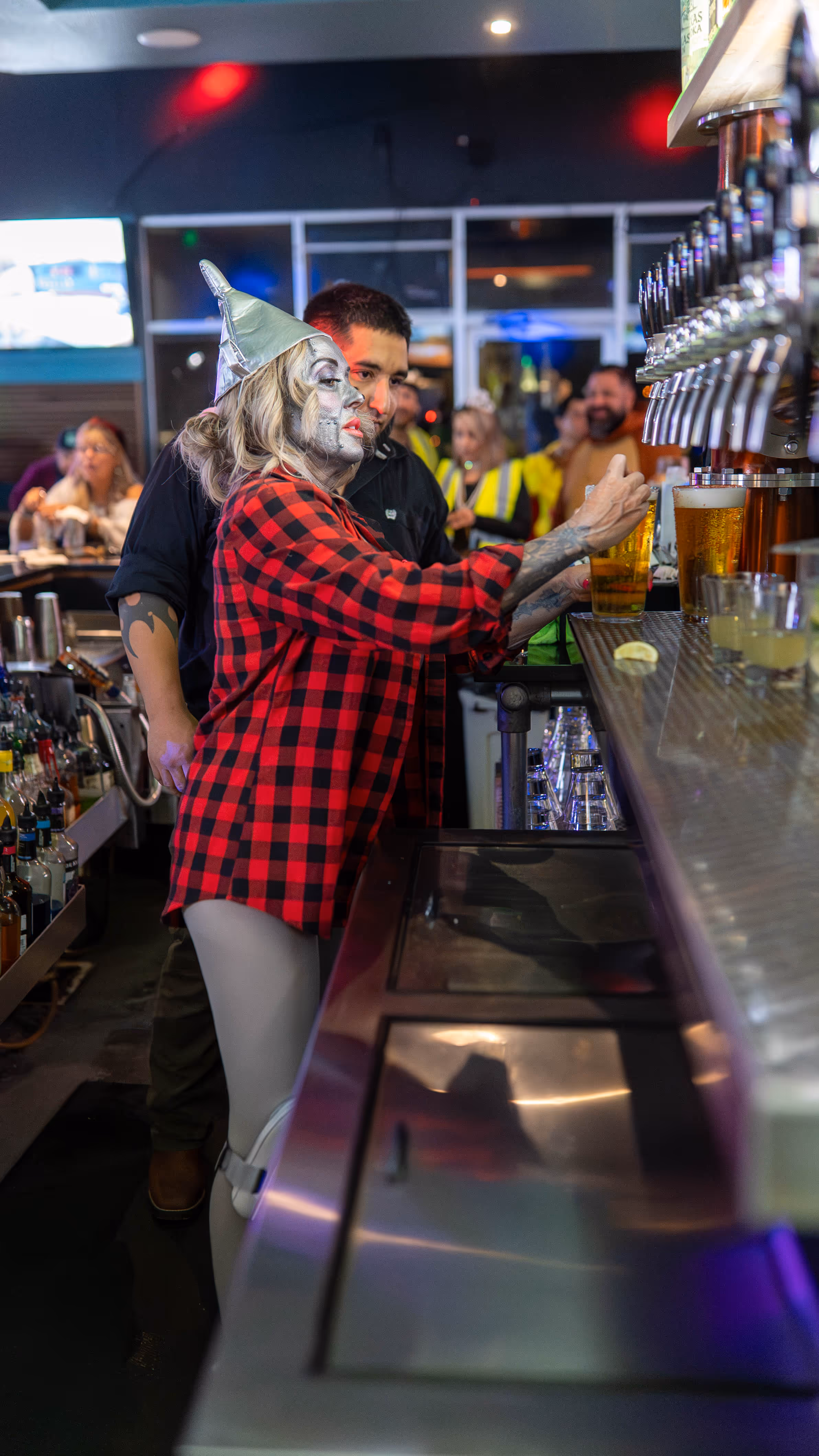 A person in silver face paint and a metallic pointed hat wearing a red and black plaid shirt is pouring a beer from a tap behind a bar.