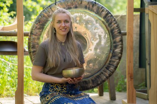 Junge Frau sitzt vor einem großen Gong und hält eine Klangschale in den Händen.