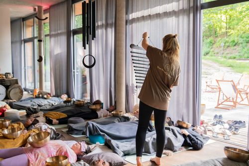 Frau spielt Klangstab vor liegenden Teilnehmern bei Klangmeditations-Workshop in hellem Raum.