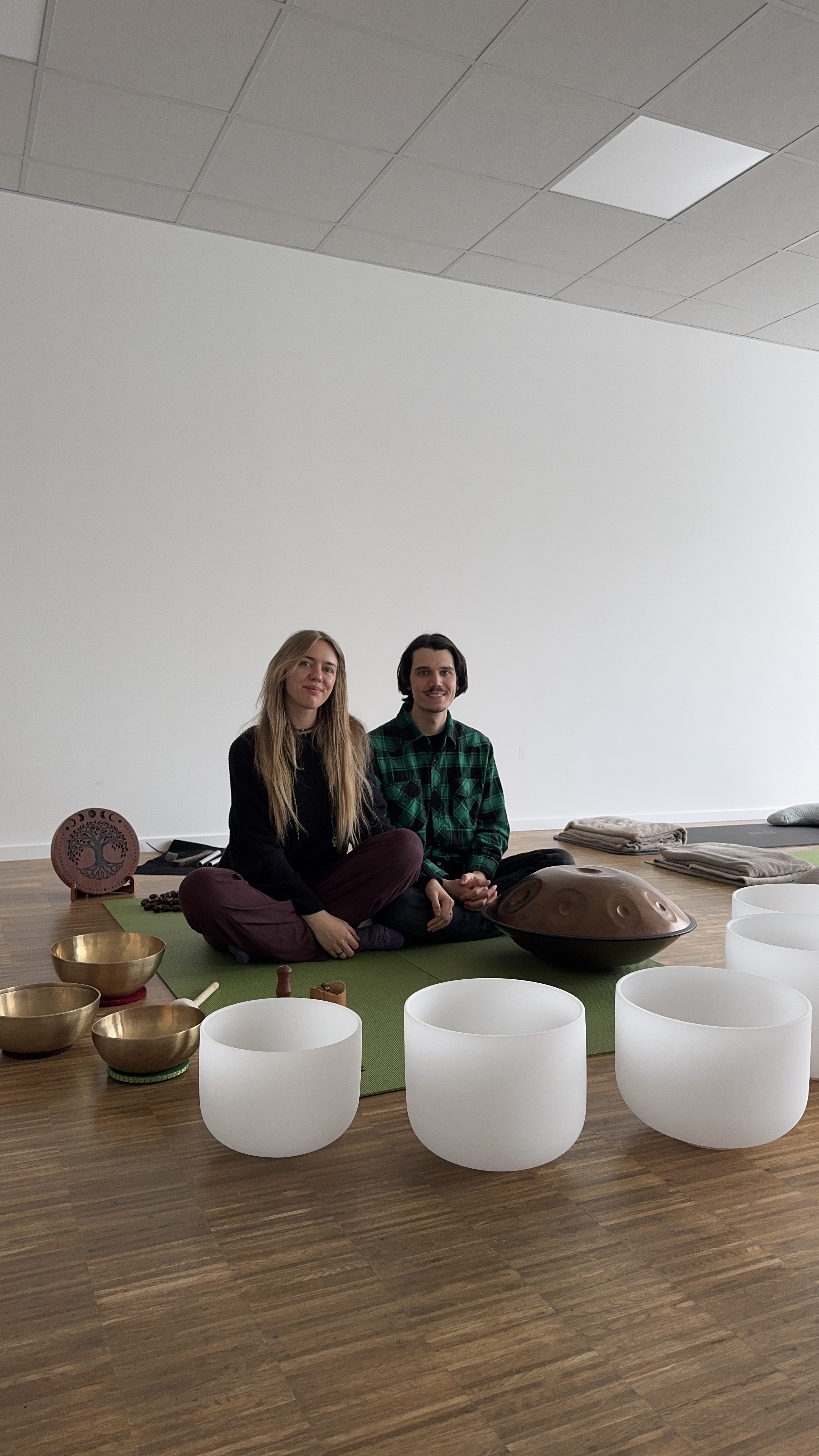 Zwei Personen sitzen auf grüner Matte mit Klangschalen und Handpan-Instrumenten in einem Raum.