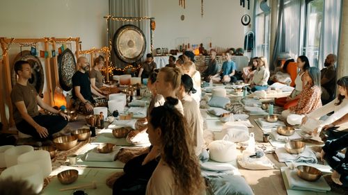 Gruppe meditierender Menschen auf Matten um Klangschalen und Gongs in einem hellen Raum.