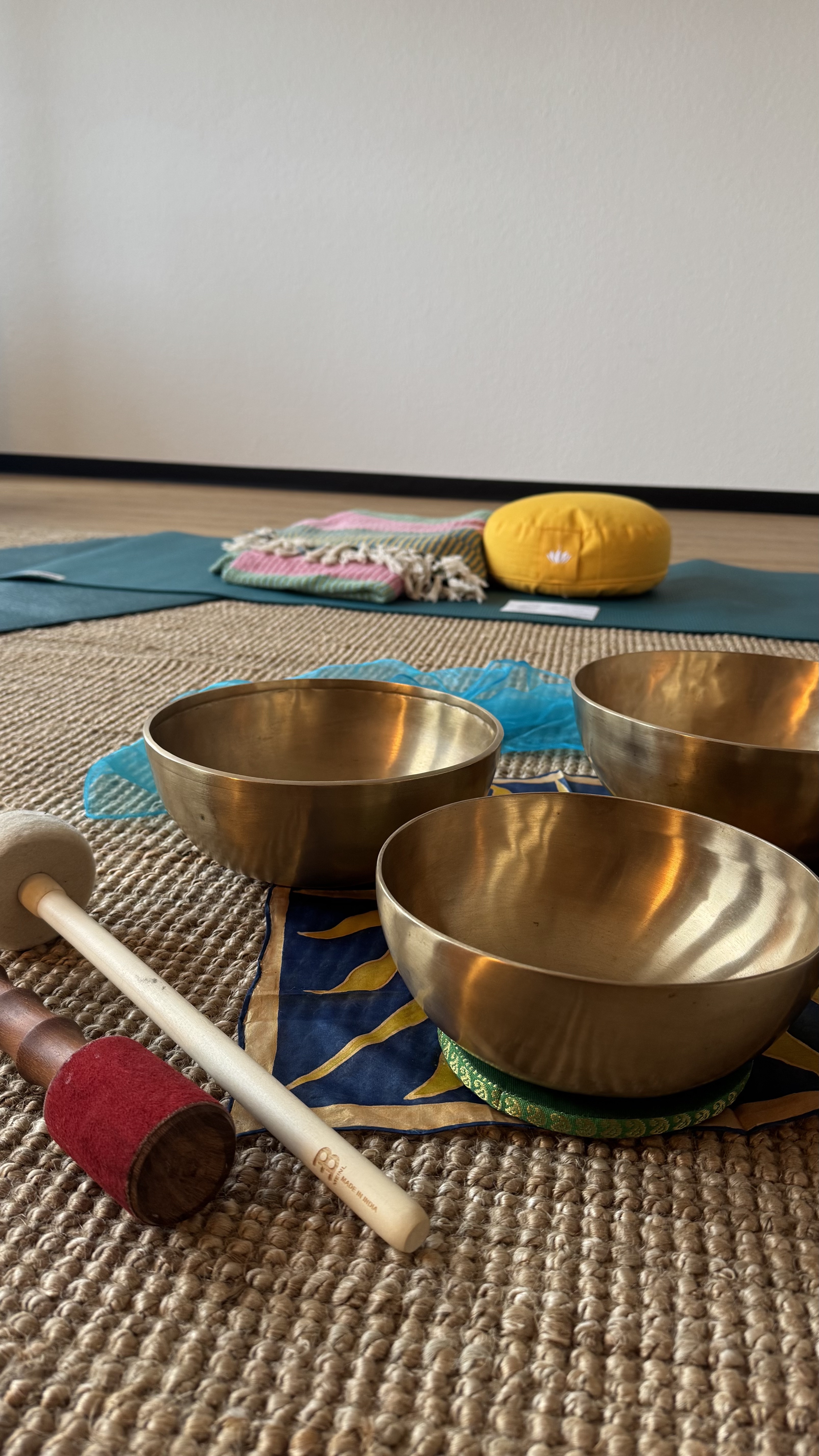 Drei Klangschalen mit Schlägeln auf einem Teppich, im Hintergrund Yogamatte, Polster und Decke.