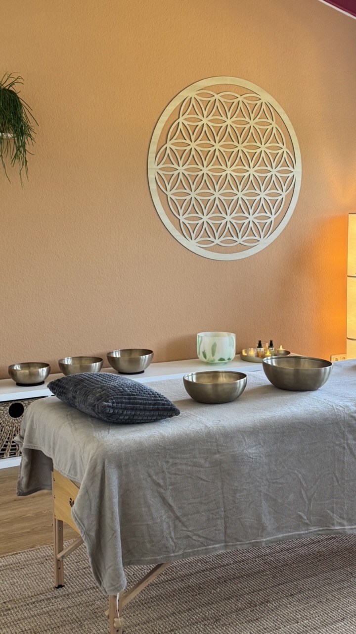 Massagebank mit Klangschalen und Kissen vor Wand mit Holzblume des Lebens Dekoration.