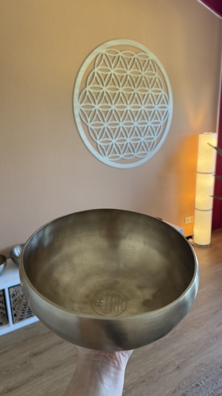 Hand hält eine bronzefarbene Klangschale vor einer Wand mit Blume des Lebens Dekor.